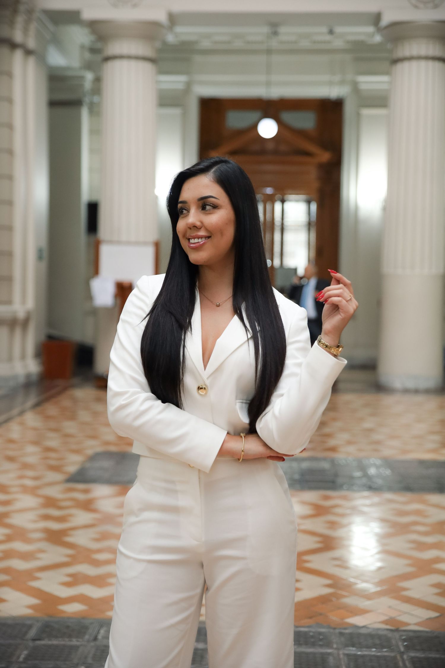 Elegant woman in white pantsuit smiling confidently inside a grand columned institutional building.