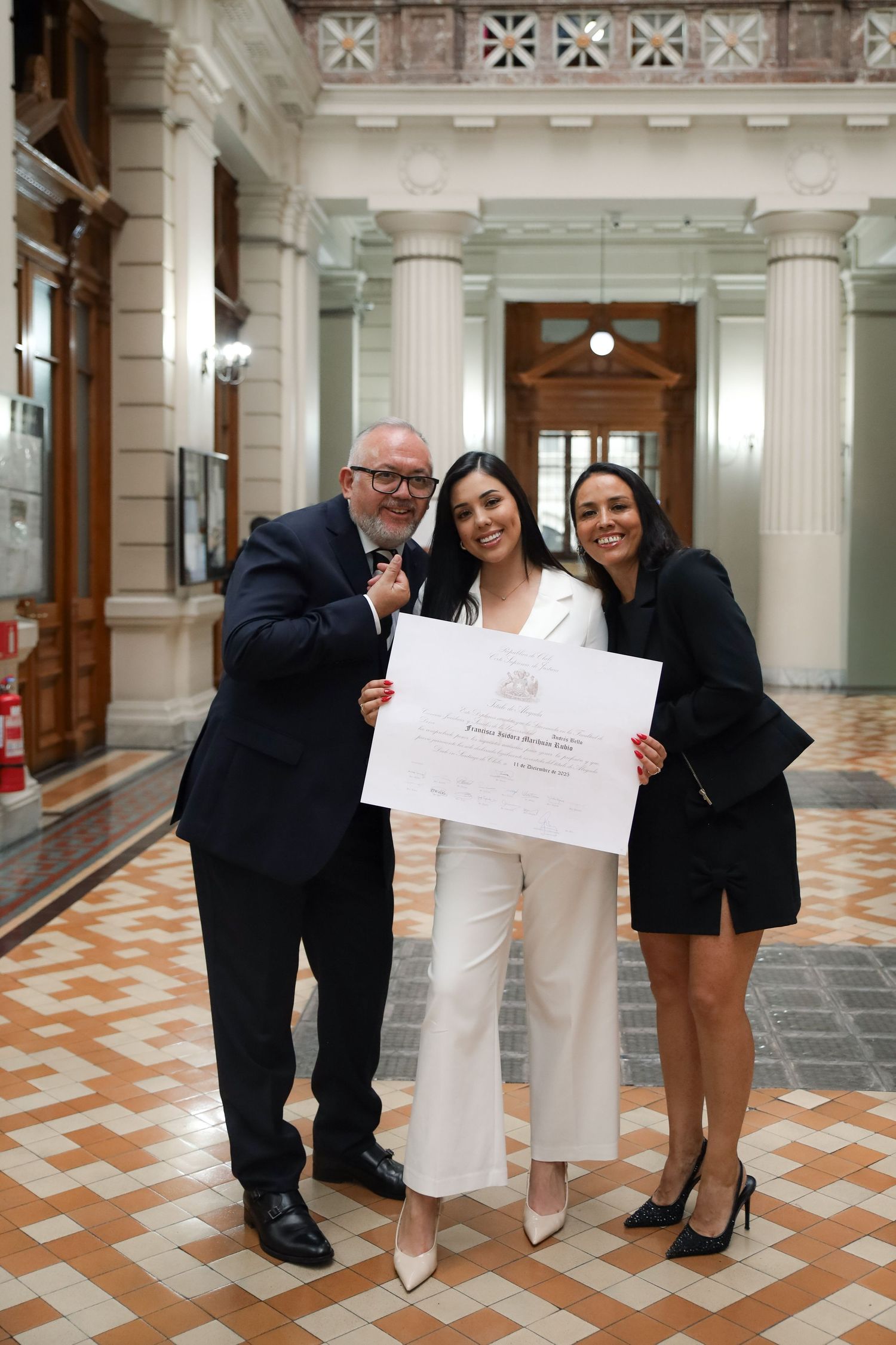 Graduate in white suit holds diploma with two proud family members in elegant columned hall.