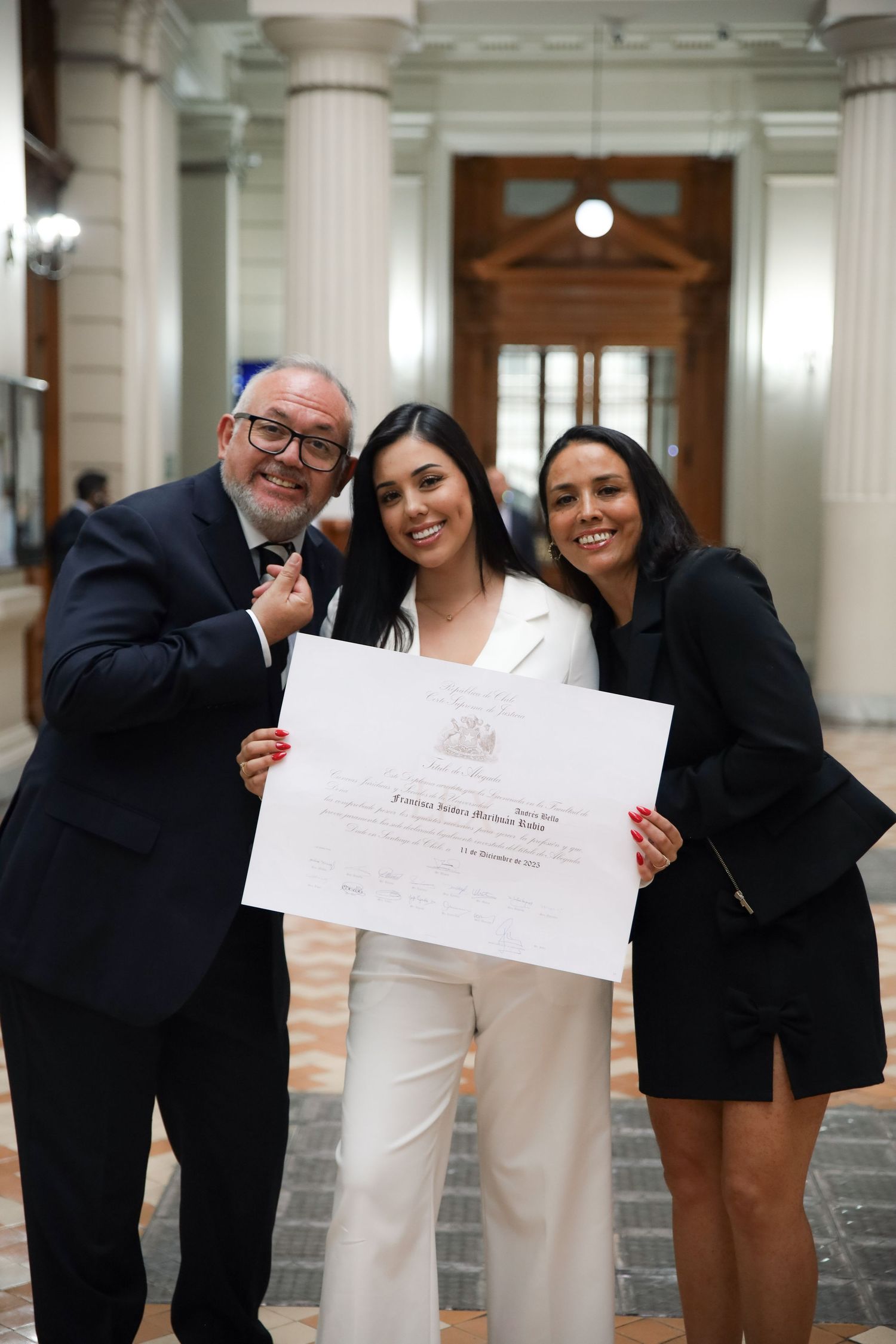 Graduate in white suit holds diploma with two proud family members in formal building.