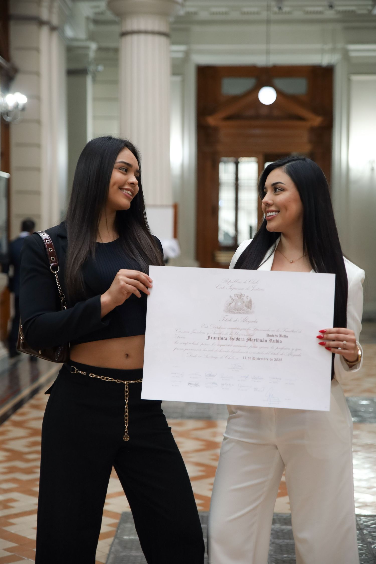 Two women in black and white outfits proudly display graduation diploma in ornate building.
