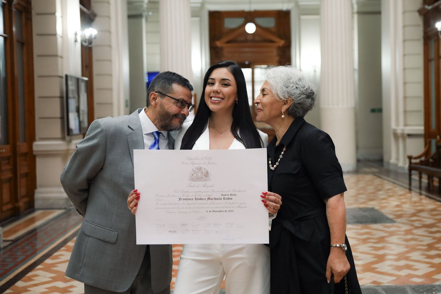 Female graduate in white suit shares joyful moment with elderly relatives holding diploma.