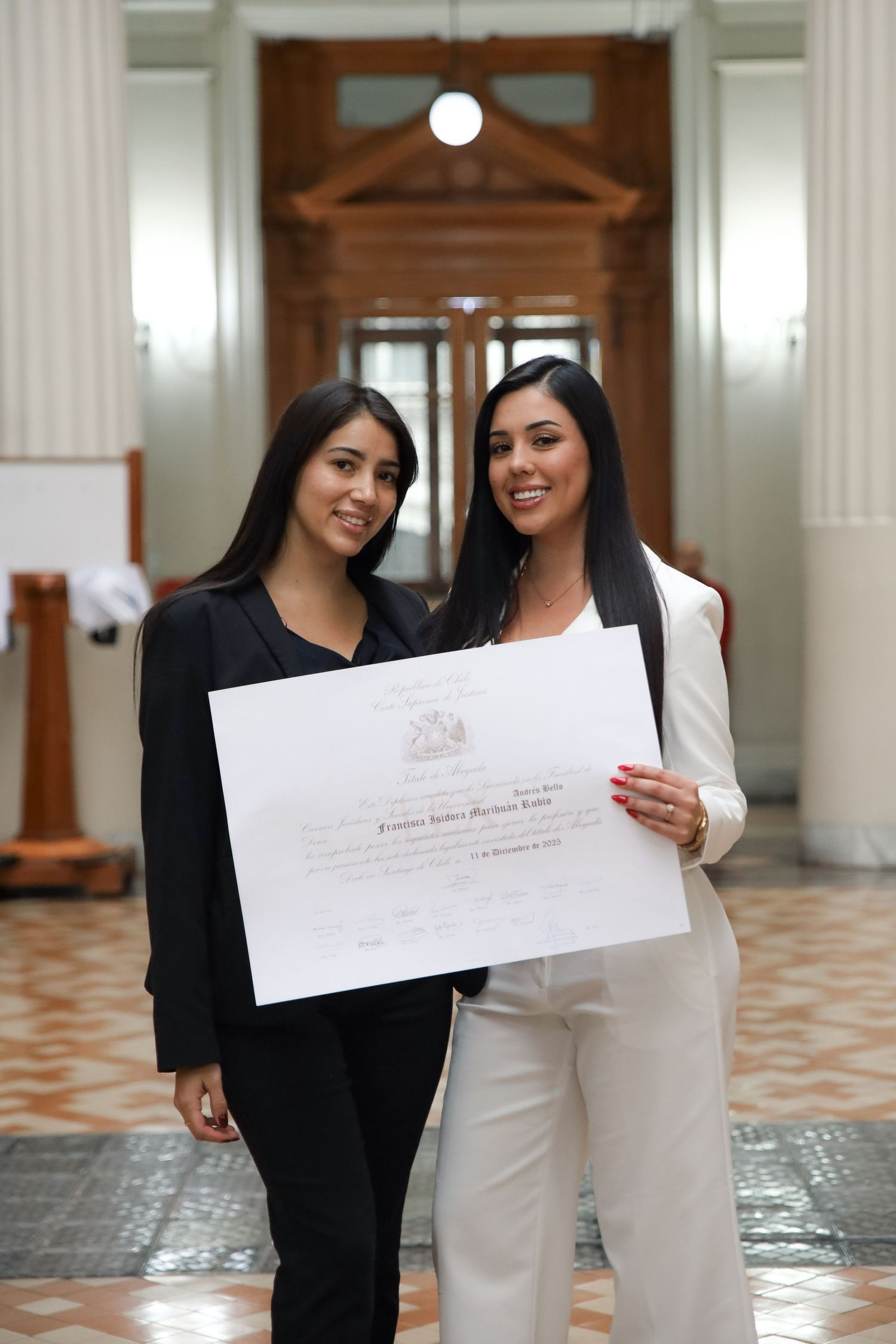 Two young women pose with graduation diploma in front of elegant columned hallway entrance.