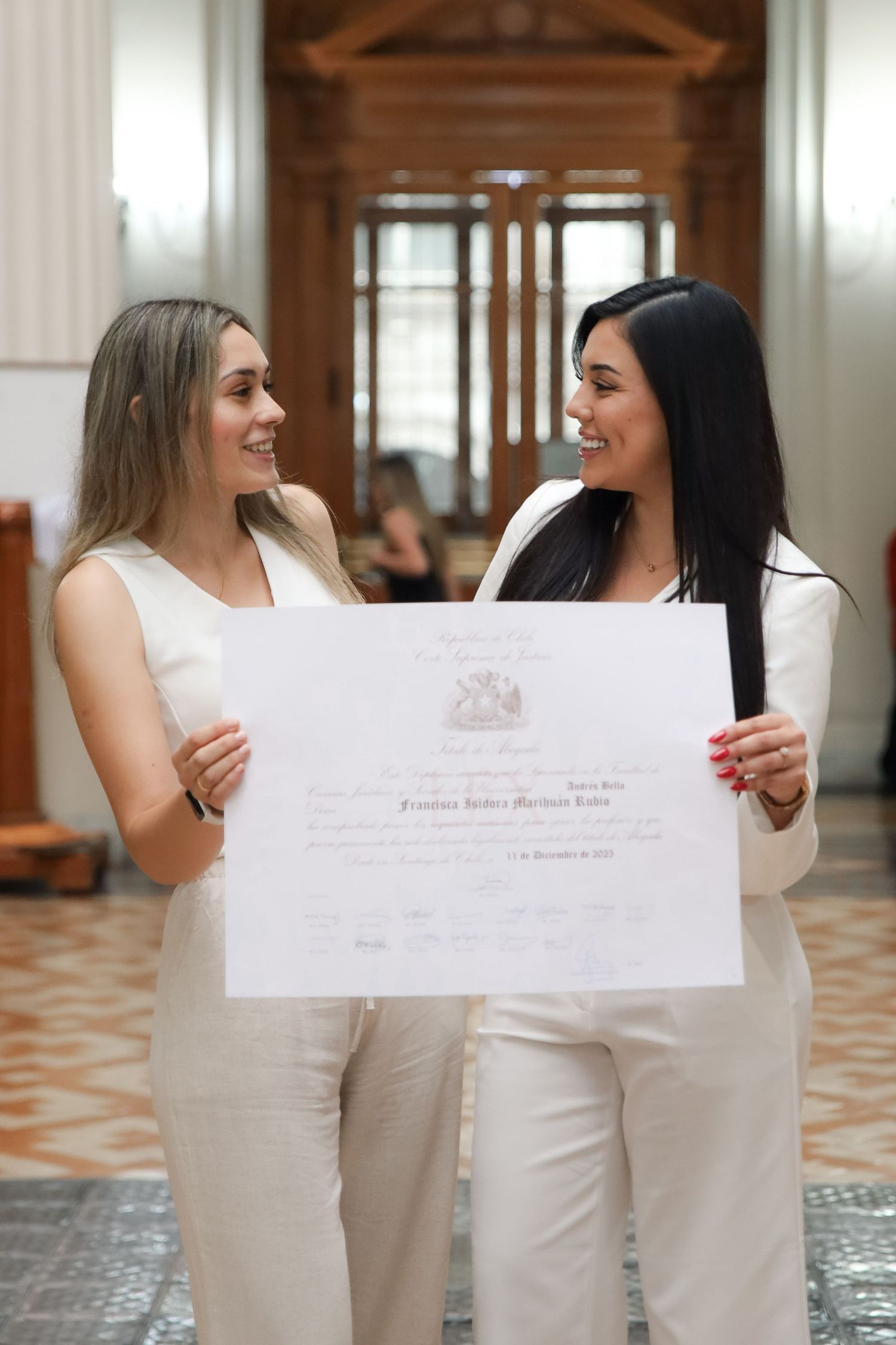 Two smiling women in white outfits hold graduation certificate in grand classical building.
