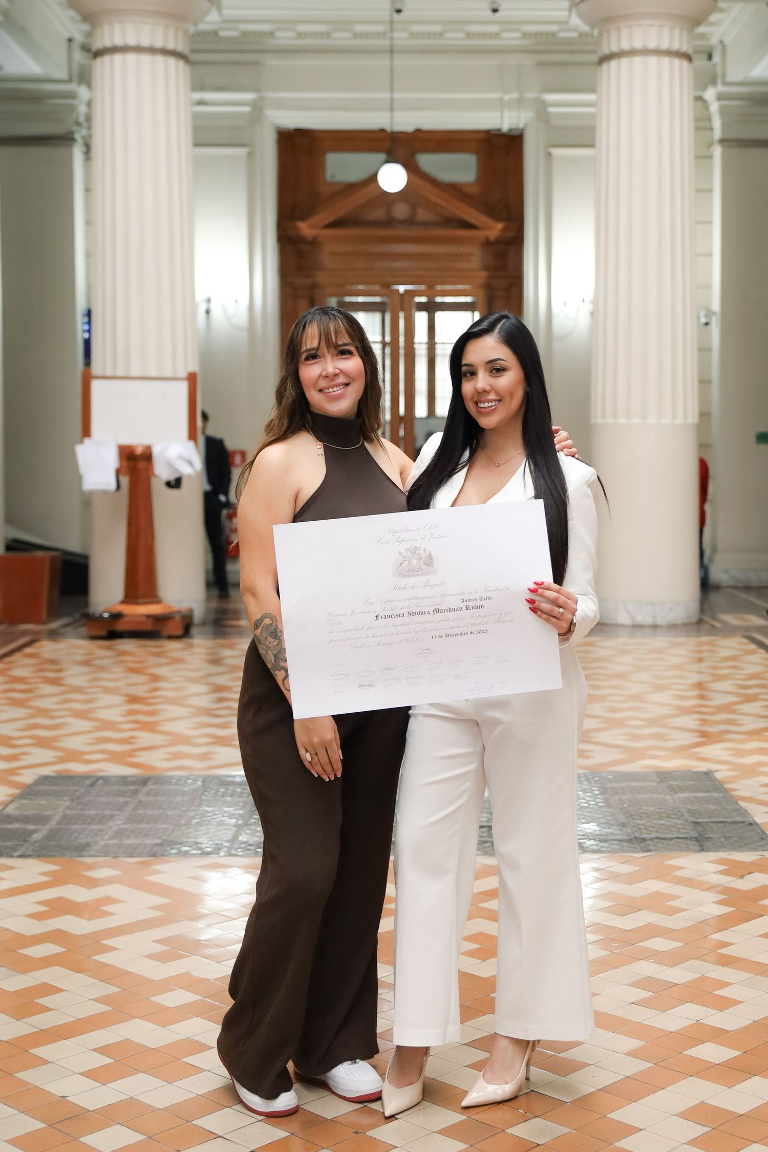 Two women proudly display graduation diploma while posing in elegant columned building interior.