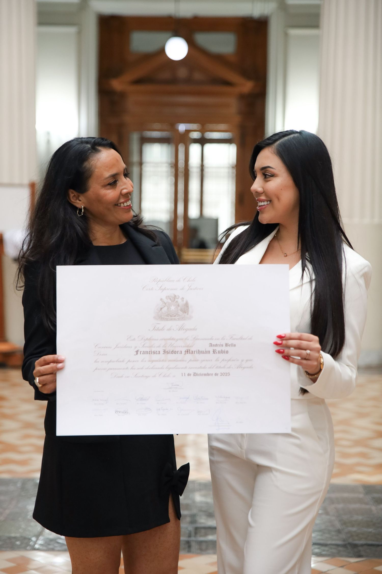 Two women smile while holding graduation diploma together in ornate classical building hallway.