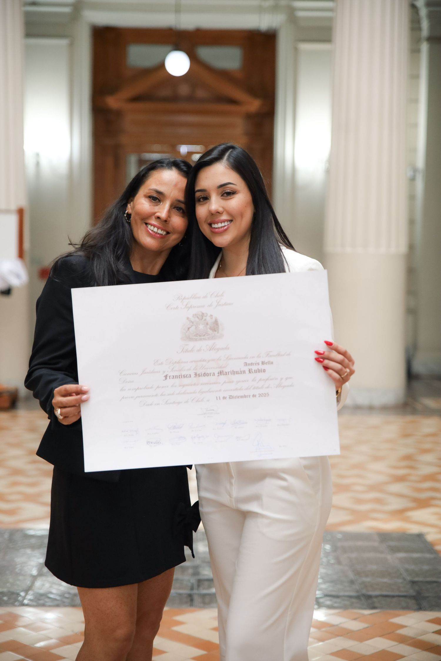 Two happy women embrace while holding graduation certificate in elegant classical building interior.