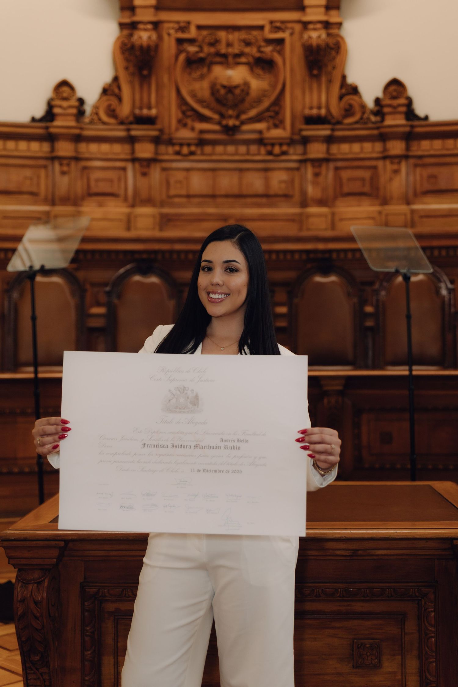Graduate in white suit proudly holds diploma in front of ornate wooden courtroom interior.