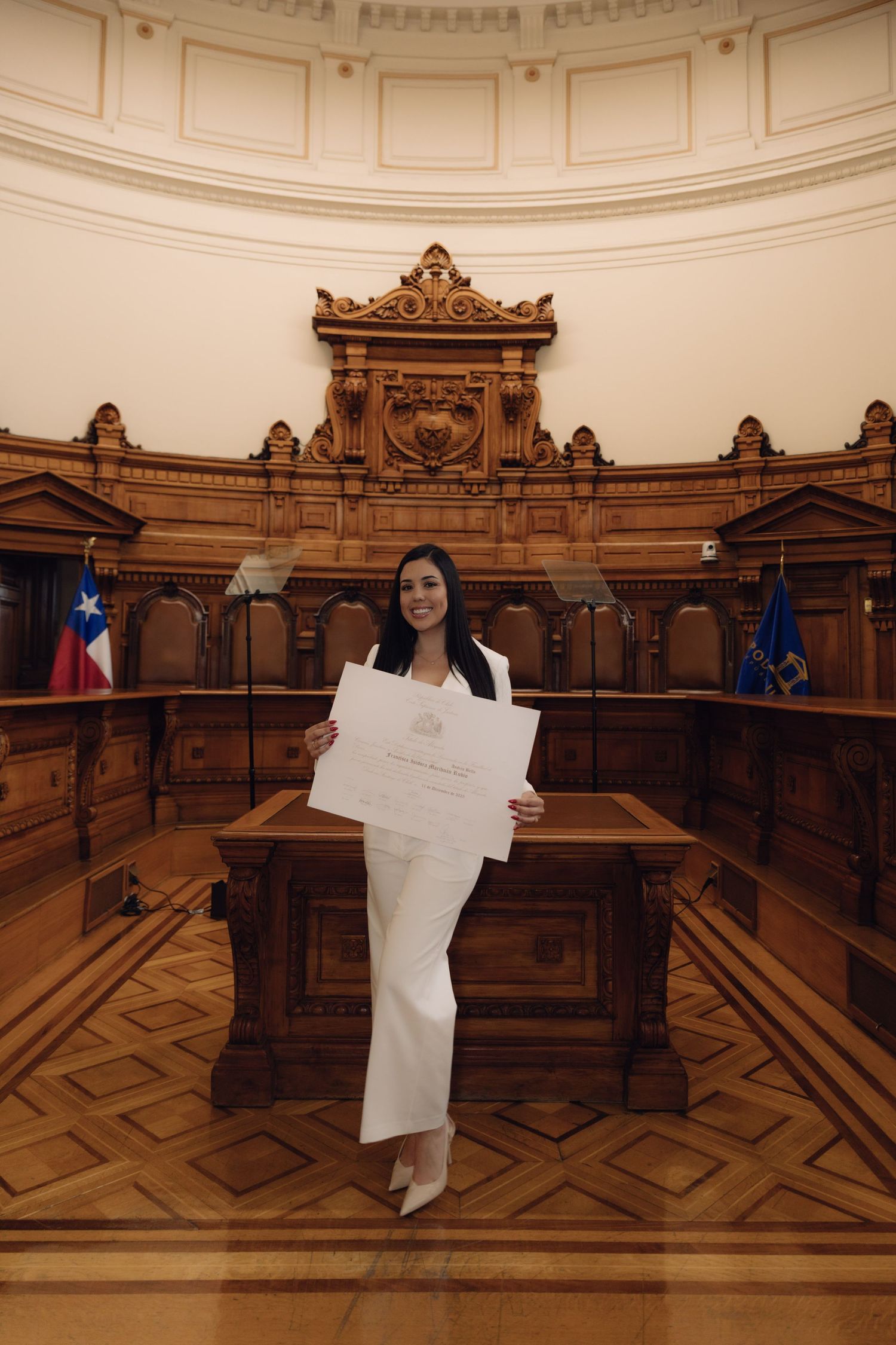 Woman in white suit holding diploma in ornate wood-paneled courtroom with Chilean flag.