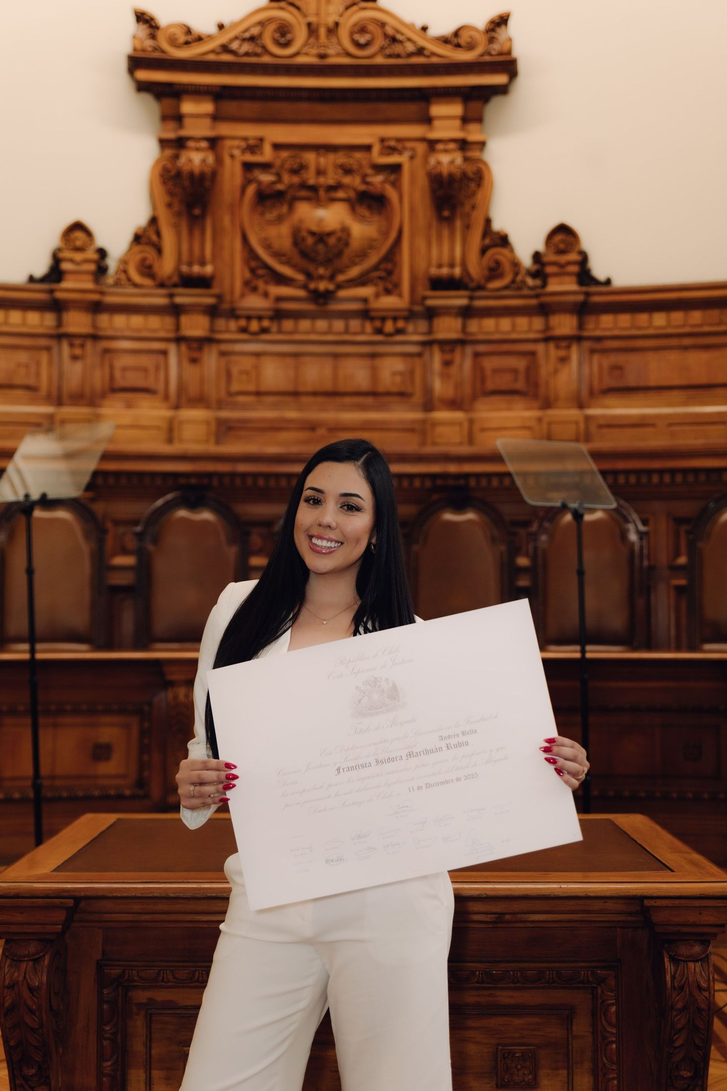 Smiling graduate in white suit proudly displays diploma in ornate wooden courtroom.