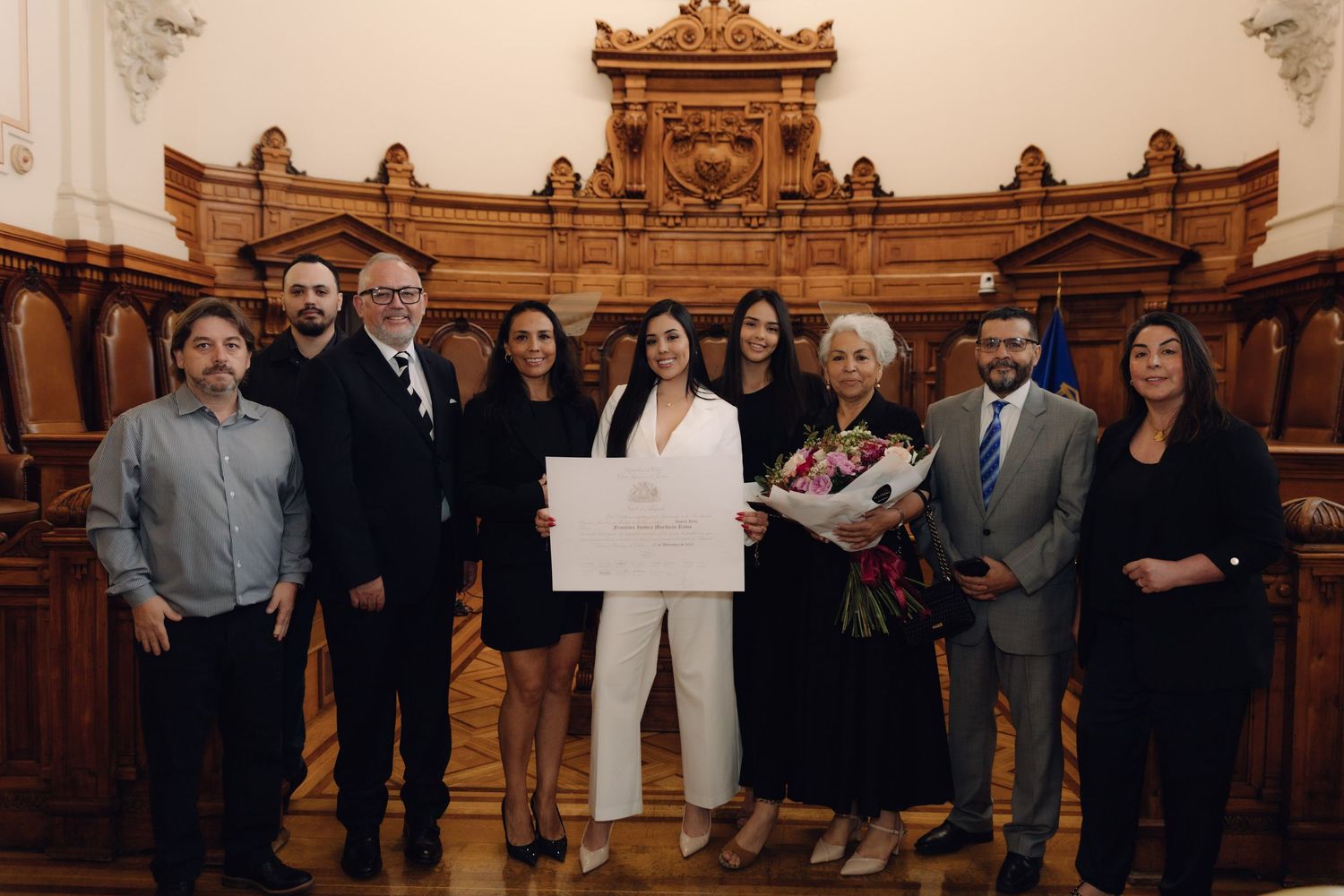 Group of nine people posing with diploma and flowers in elegant wood-paneled courtroom.
