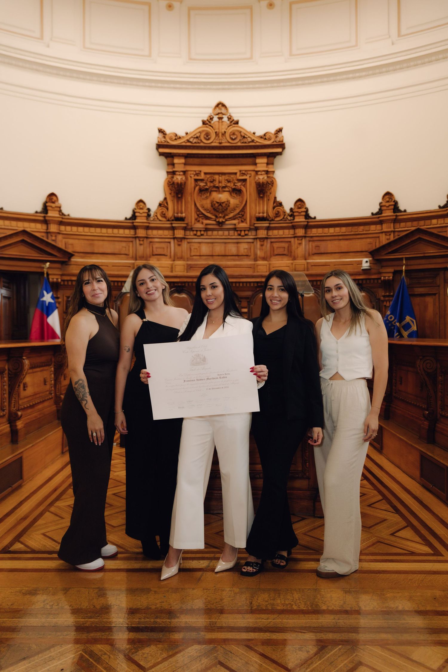 Female graduate in white suit poses with four friends holding diploma in courtroom.