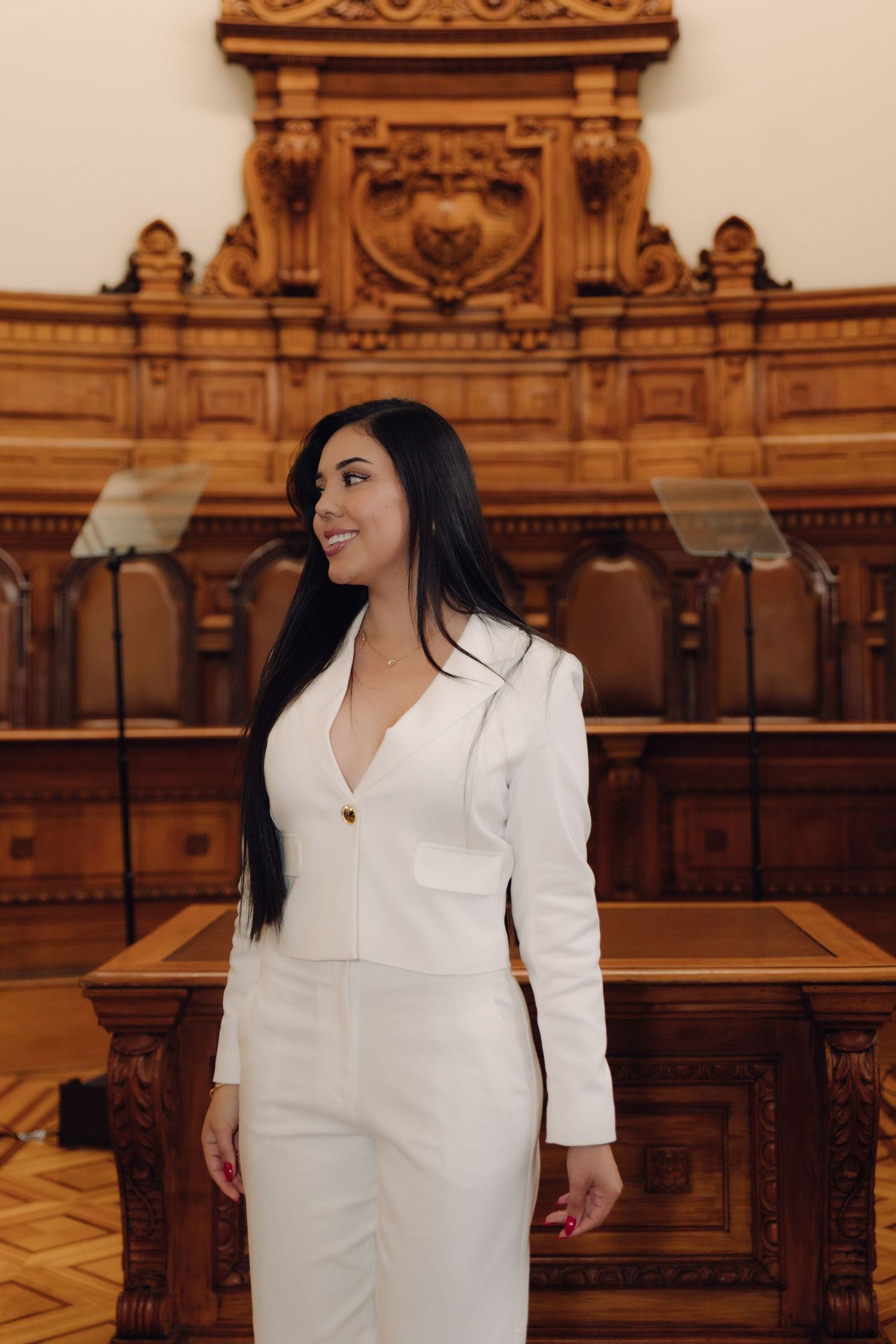 Elegant woman in white blazer smiles in ornate wooden courtroom interior.