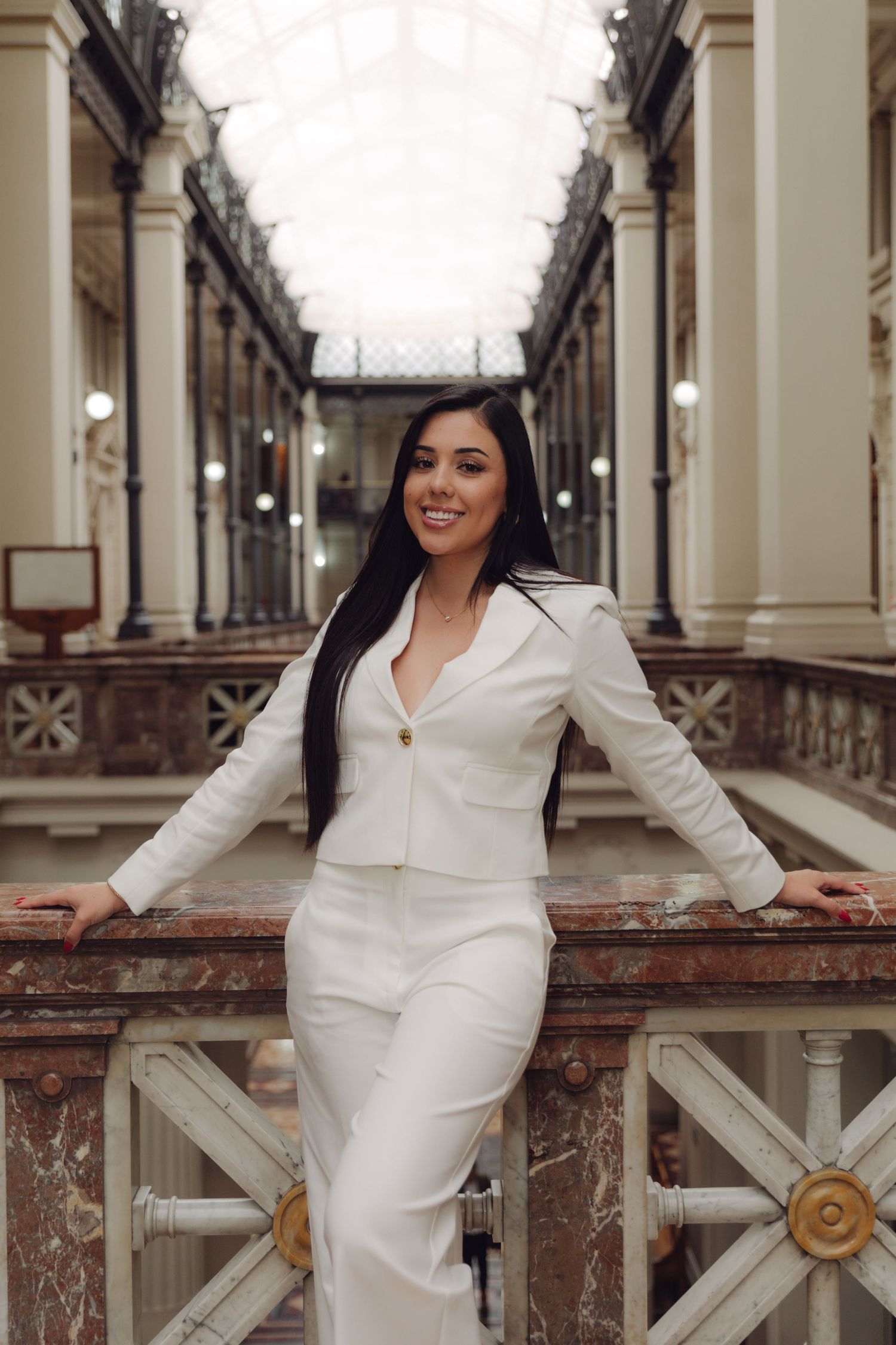 Smiling woman in white blazer leaning on marble railing in grand architectural gallery.