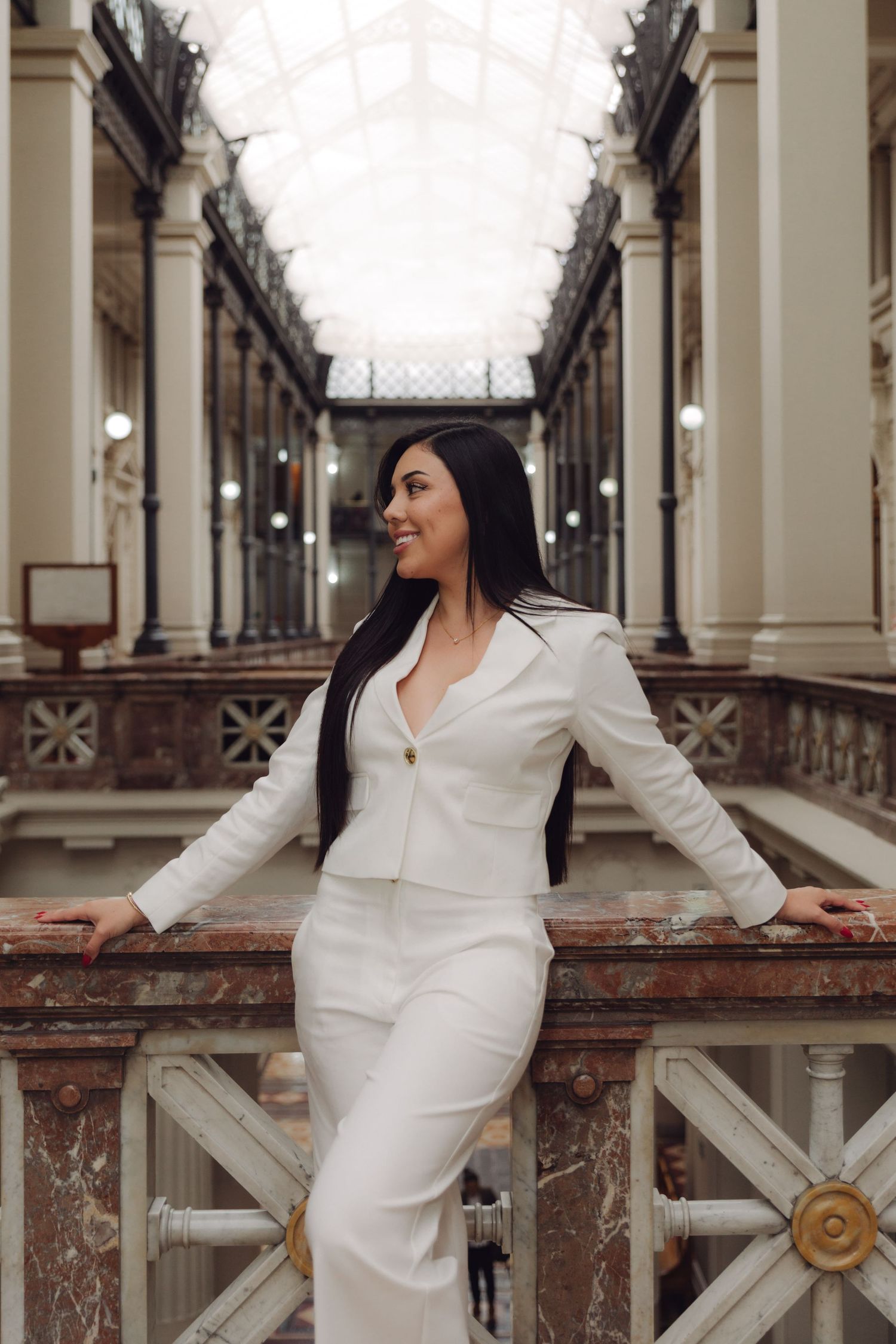 Elegant woman in white suit smiling in historic columned gallery with glass ceiling.