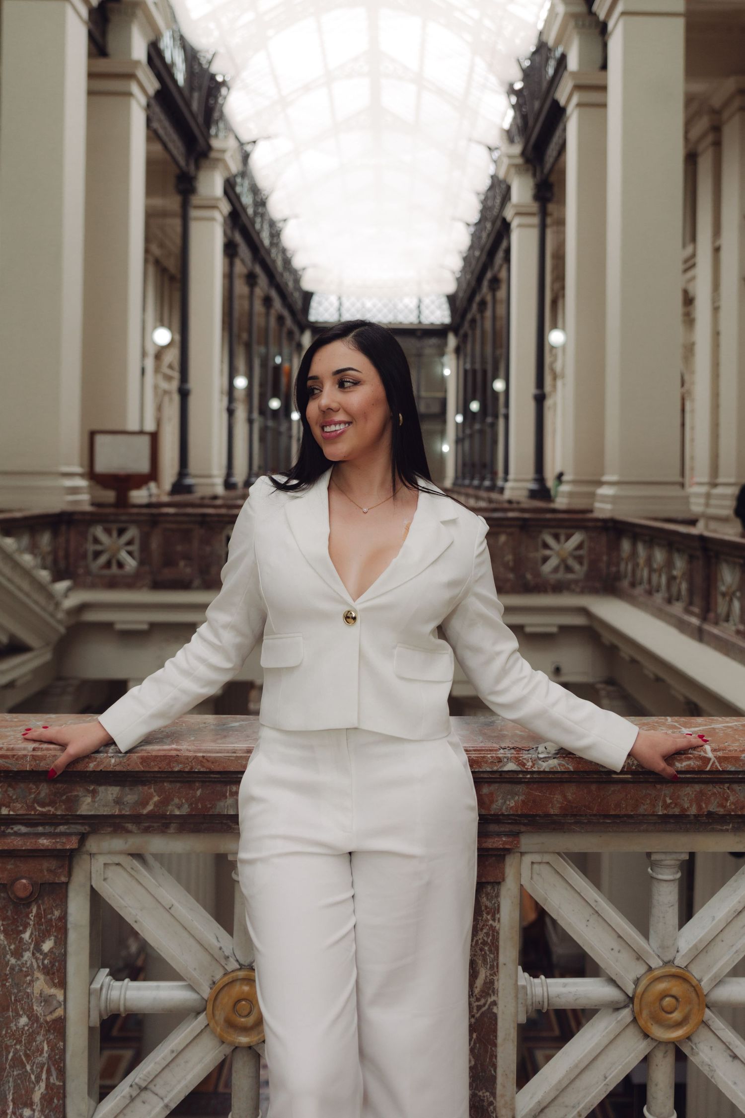 Stylish woman in white suit leans on railing in grand historic gallery corridor.