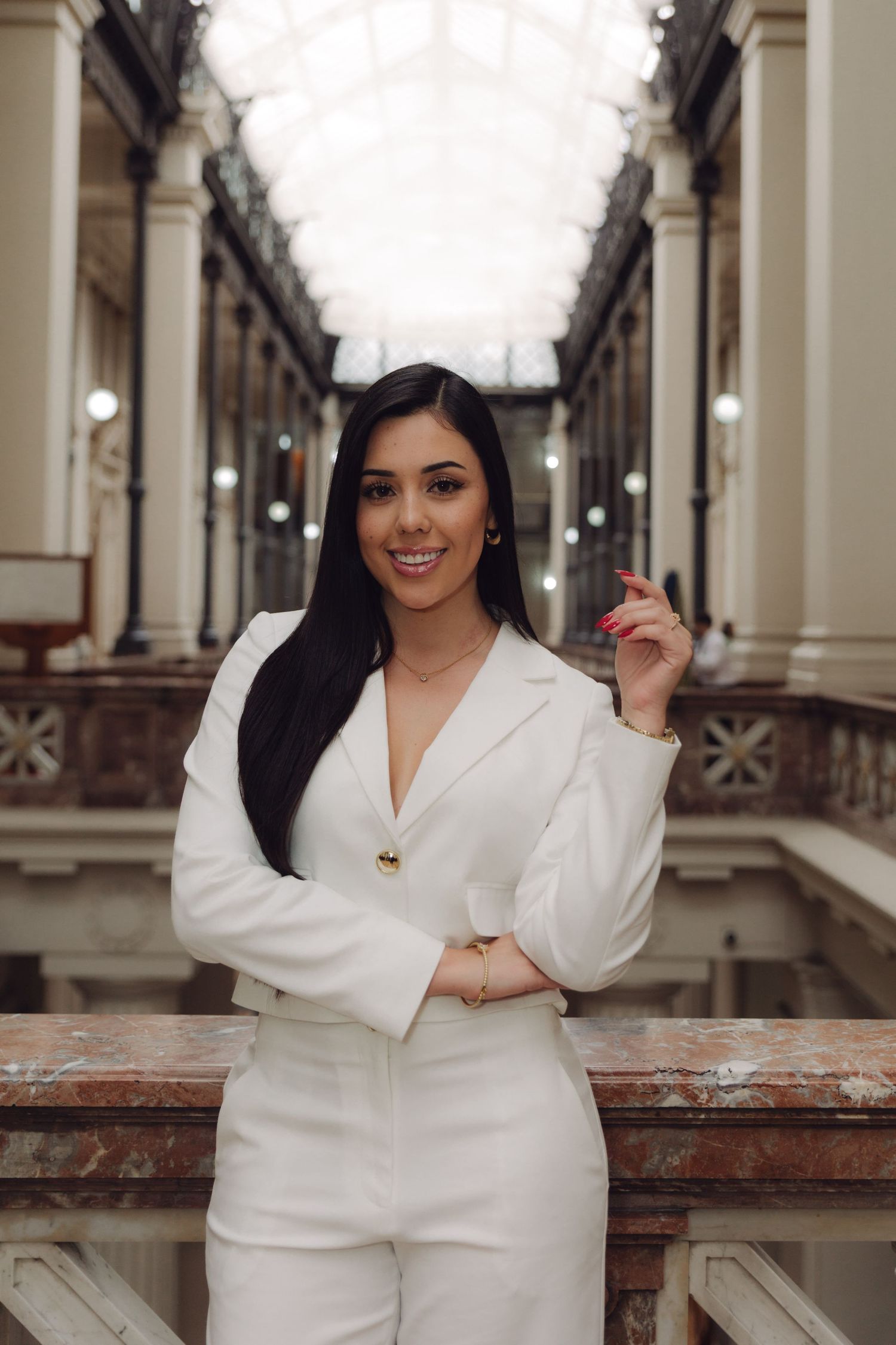 Confident woman in white blazer posing in ornate historic building interior gallery.