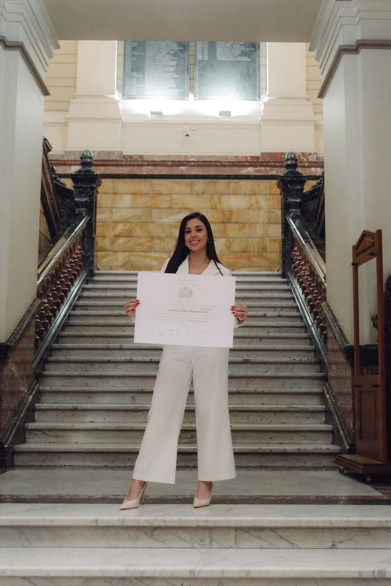 Graduate in white outfit holds diploma while standing before grand marble staircase.