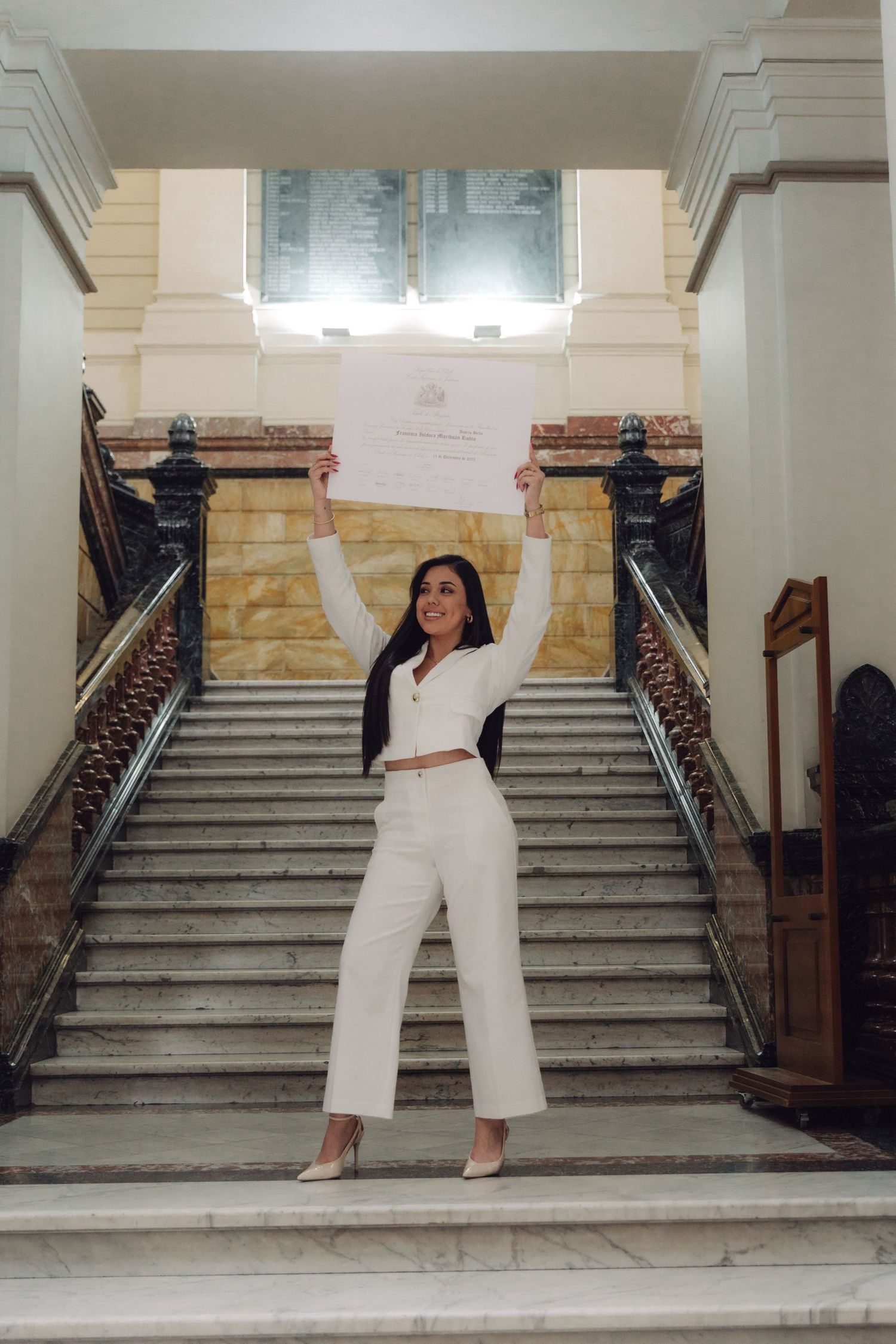 Celebrating graduate in white suit raises diploma triumphantly on elegant marble staircase.