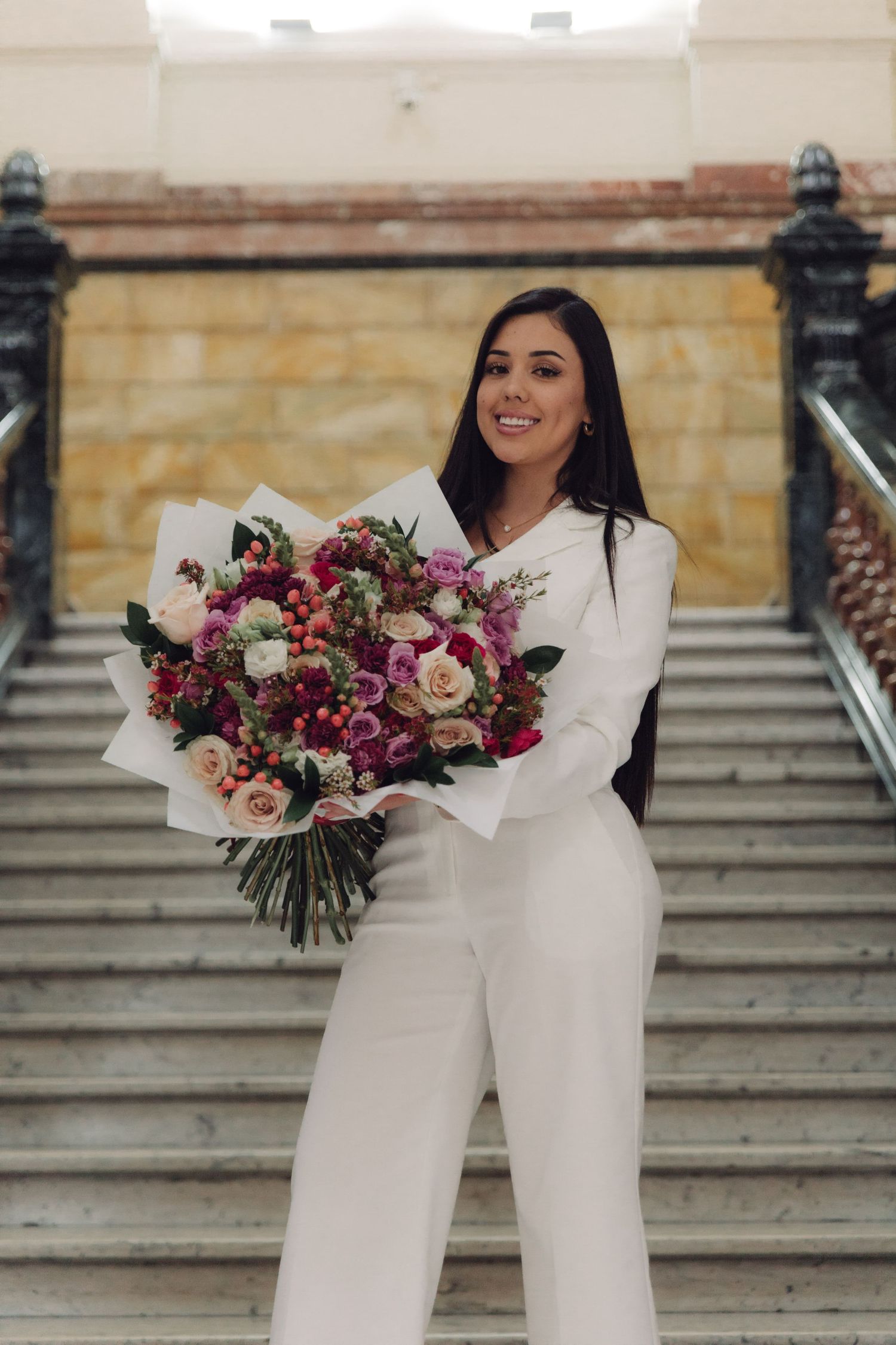 Smiling woman in white suit holding large bouquet of pink and red roses on grand staircase.