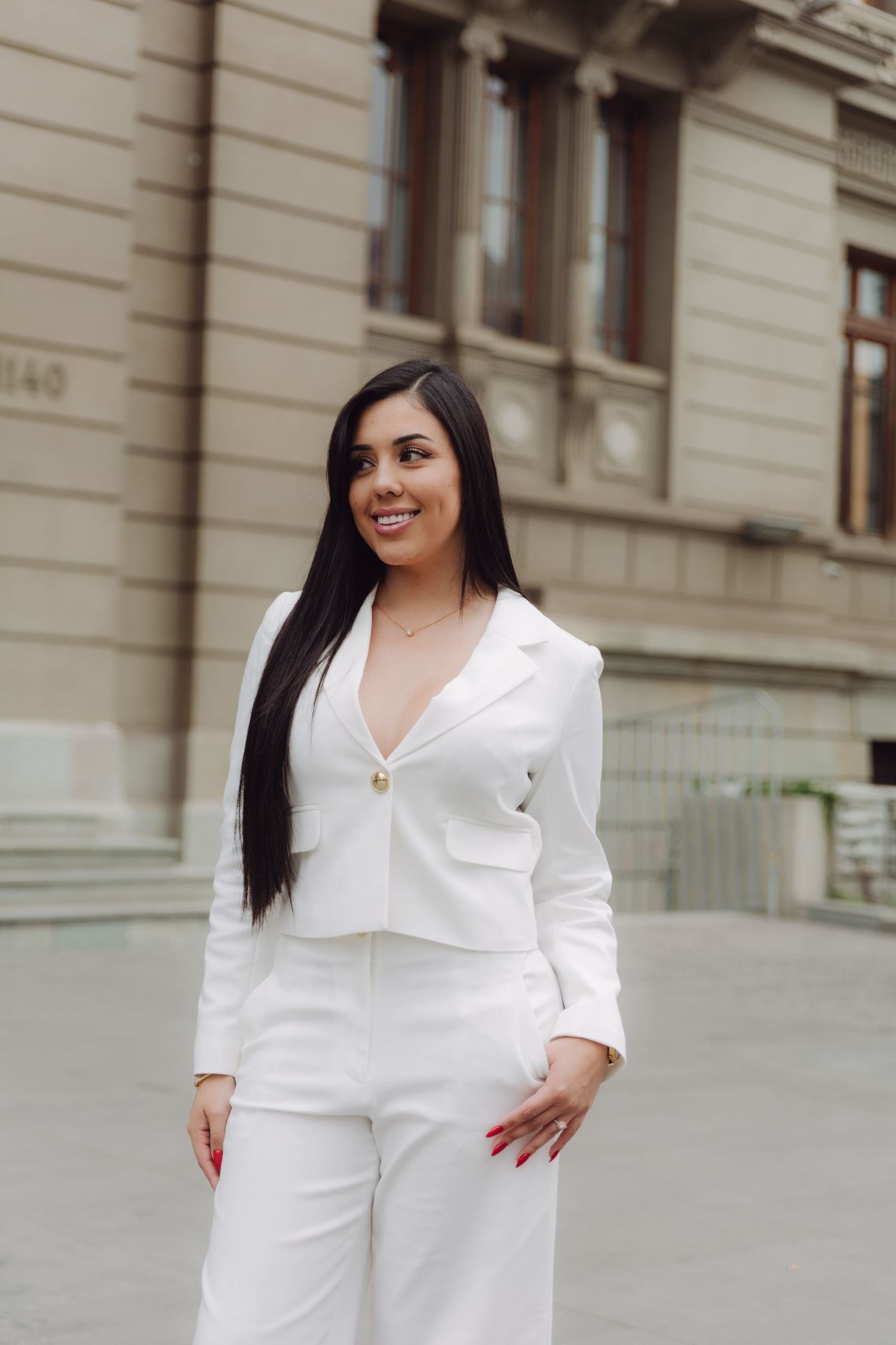 Woman in white blazer and pants smiling outdoors beside classic stone building facade.
