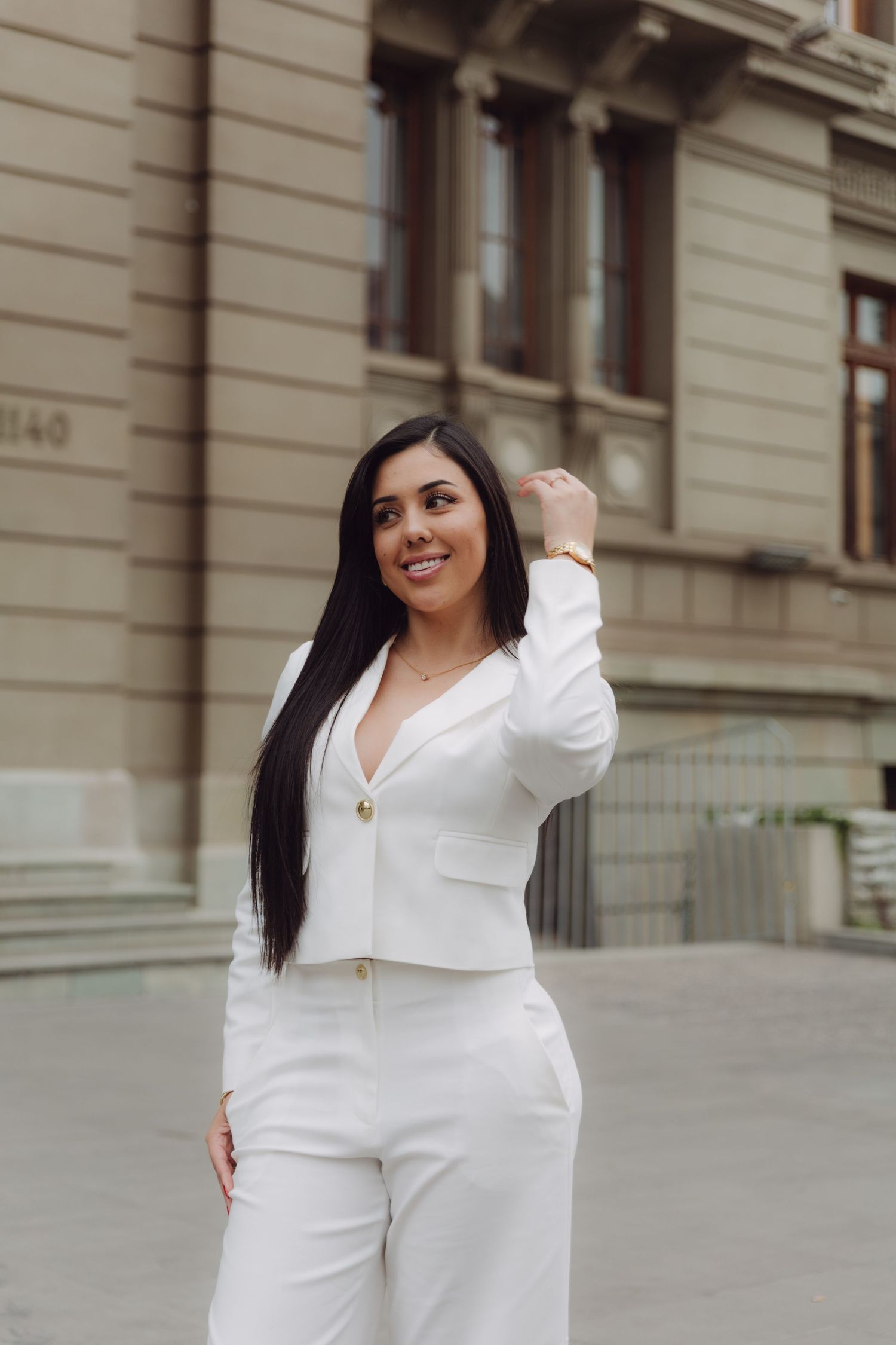 Happy woman in white suit posing outdoors near historic stone building raising hand.