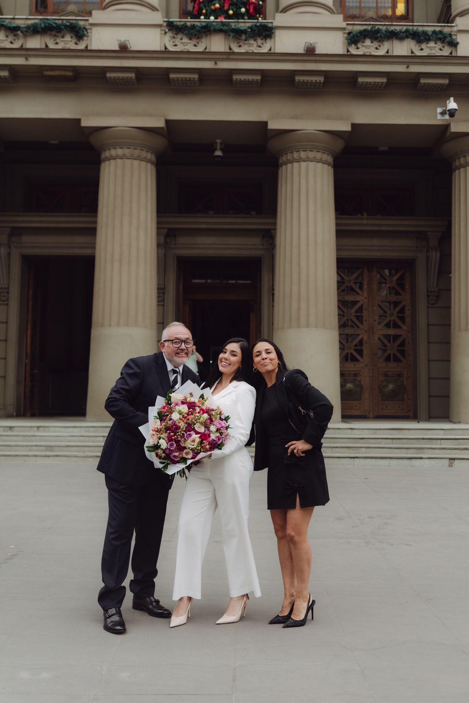Three people posing outside neoclassical building, woman in white holding colorful flower bouquet.