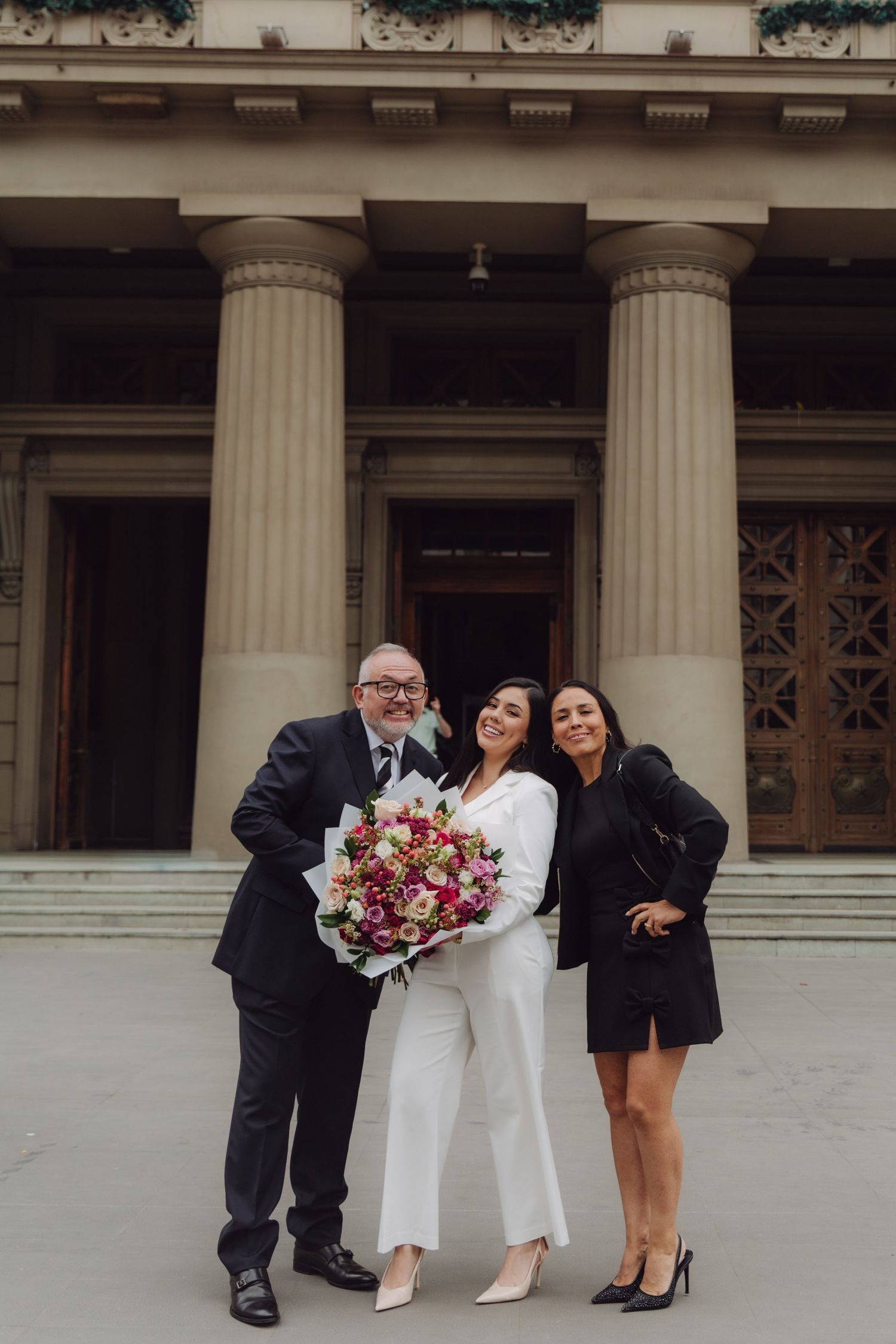 Three people posing with colorful flower bouquet outside neoclassical columned building.