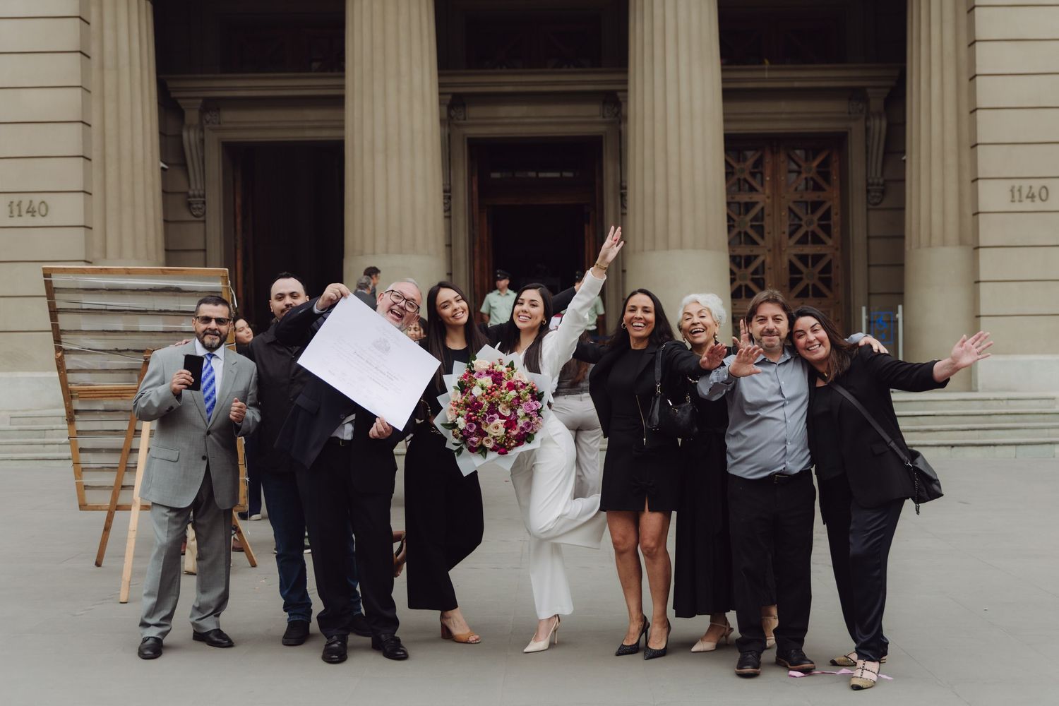 Group of people celebrating outdoors in front of classic building, woman holding bouquet and document.