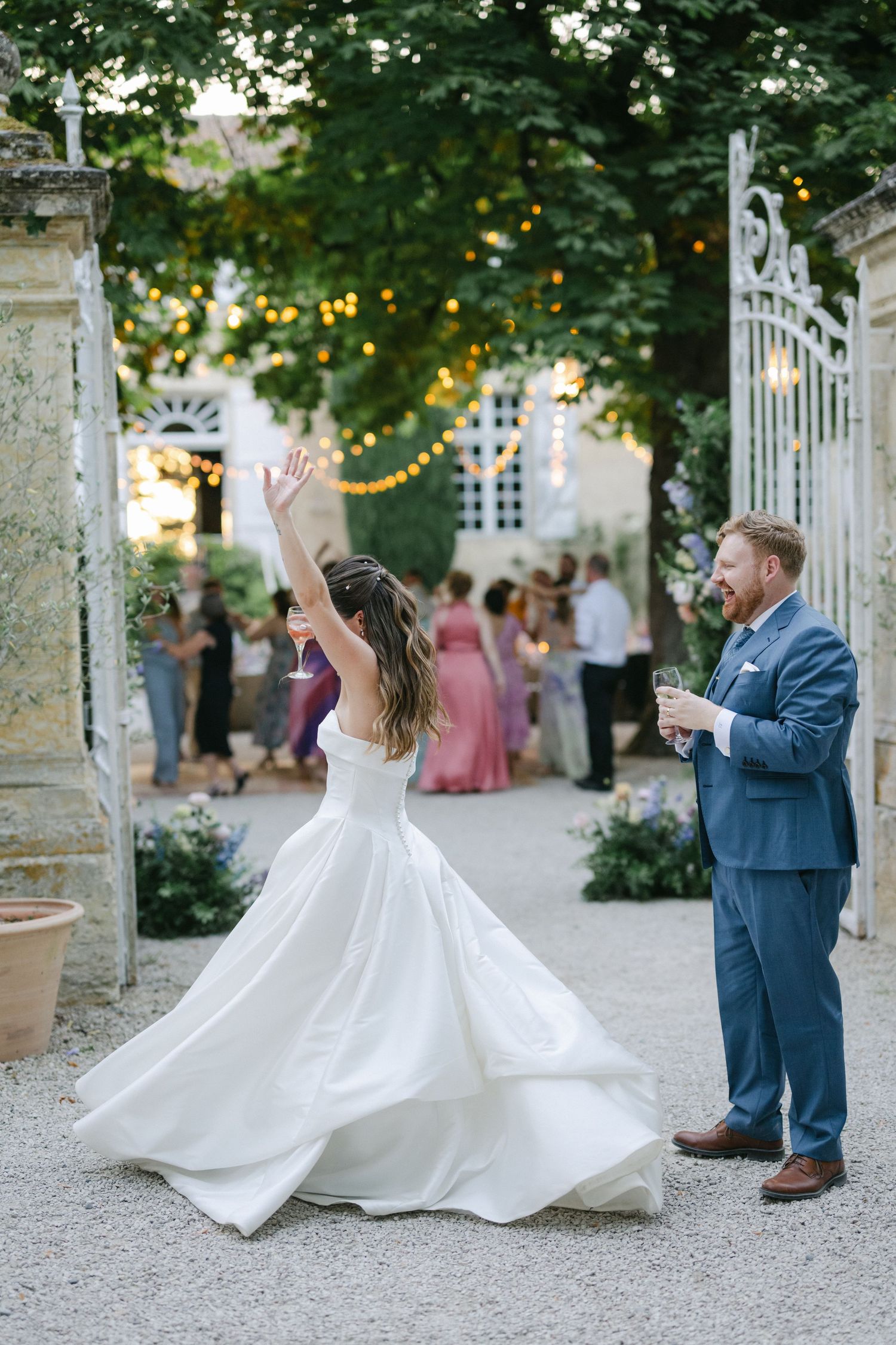 Smiling bride in a flowing white strapless gown holds a pastel bouquet outside an elegant French chateau.