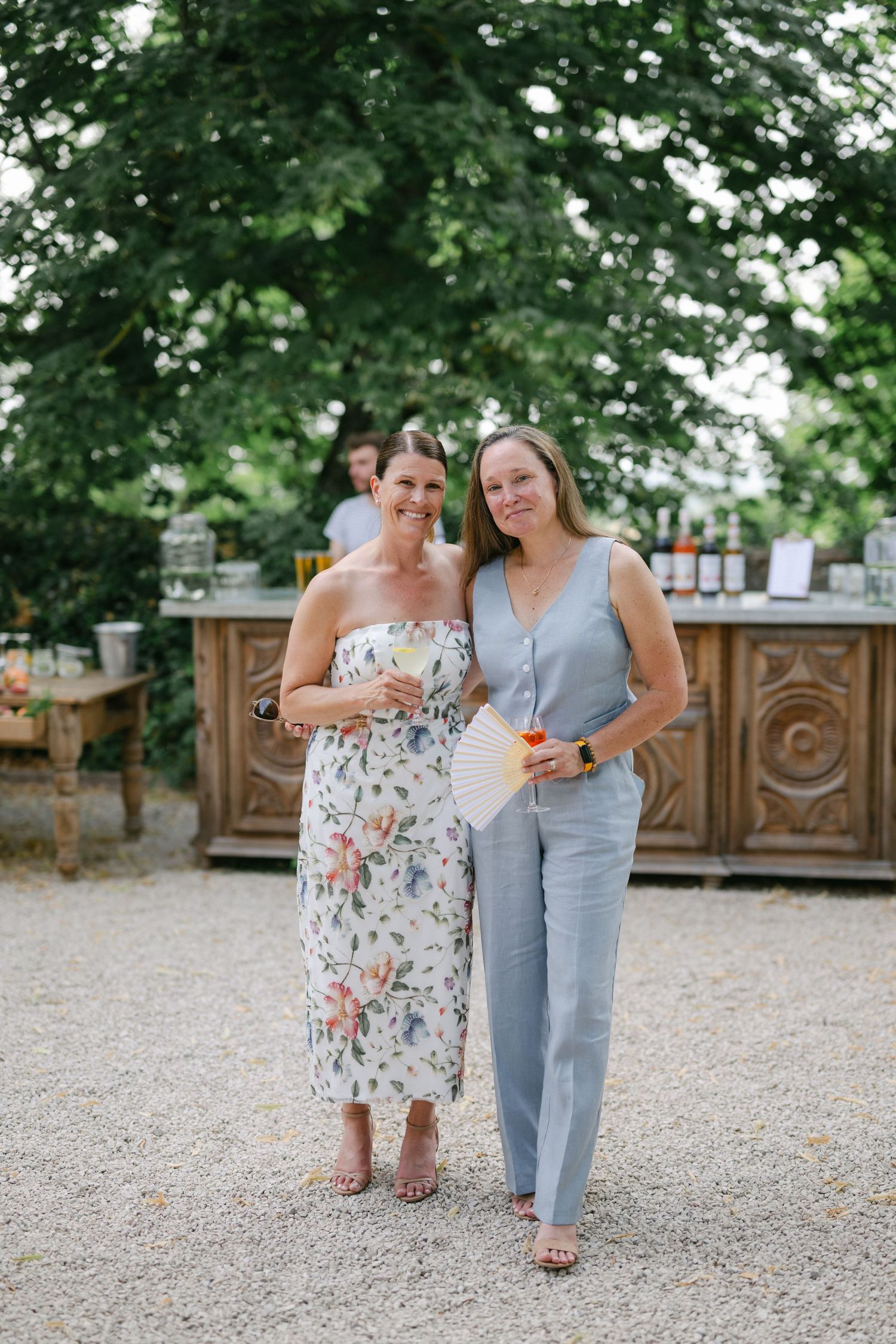 Two women in summer attire smile together at an outdoor garden party near a rustic wooden bar.