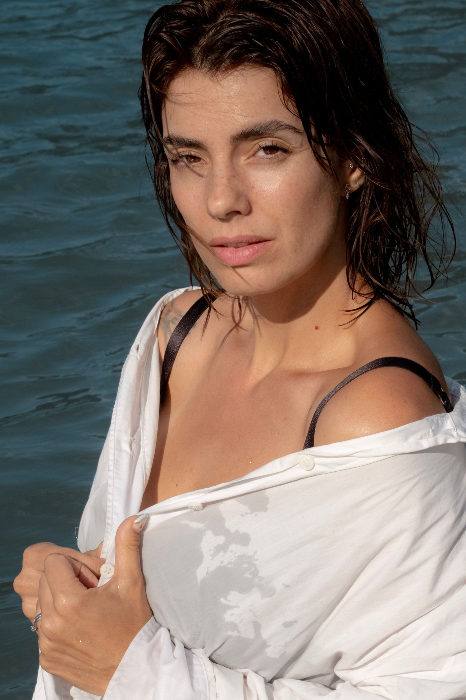 Woman with wet dark hair wearing a white shirt near the ocean, gazing at the camera.