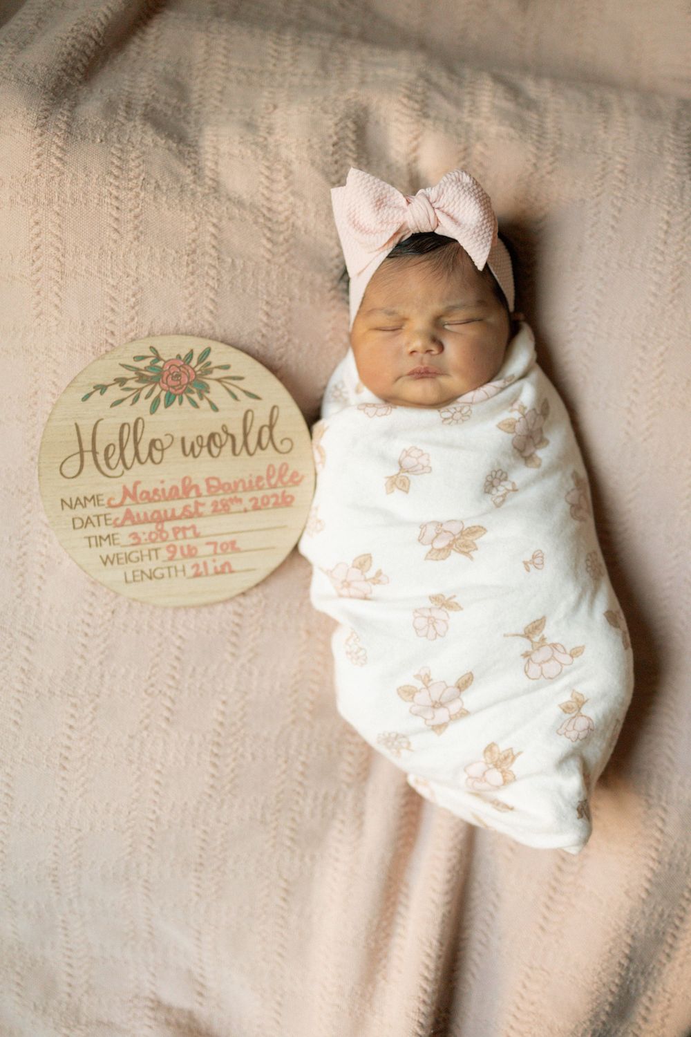 Sleeping newborn baby girl swaddled in floral wrap with pink bow headband next to Hello World birth announcement sign.