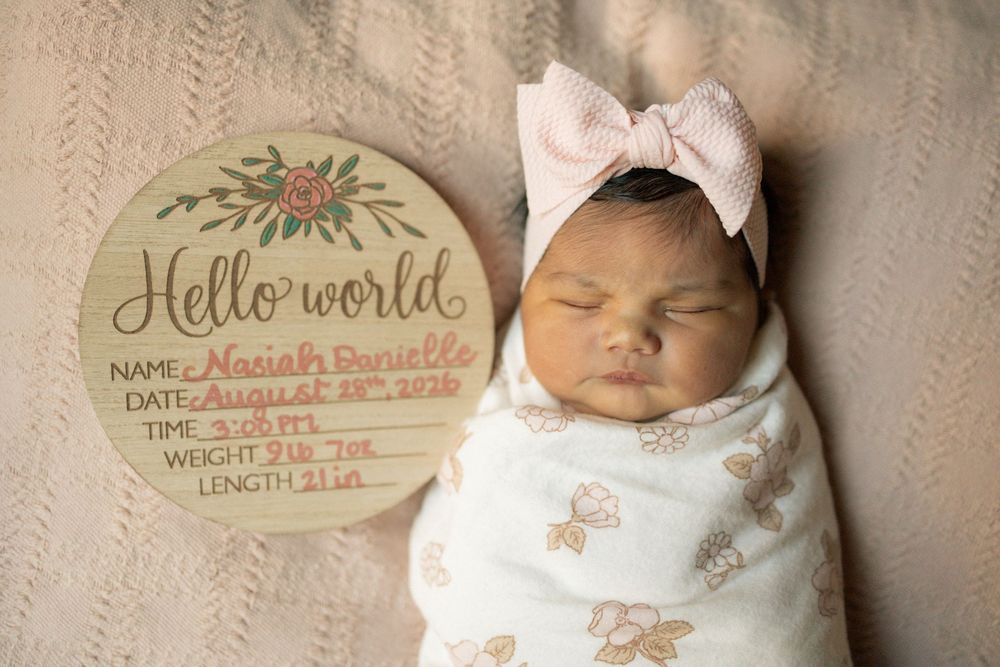 Sleeping newborn baby girl in floral swaddle with pink bow headband beside Hello World wooden birth announcement sign.