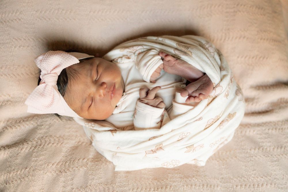 Newborn baby swaddled in white wrap with pink bow headband sleeping peacefully on a soft beige blanket.