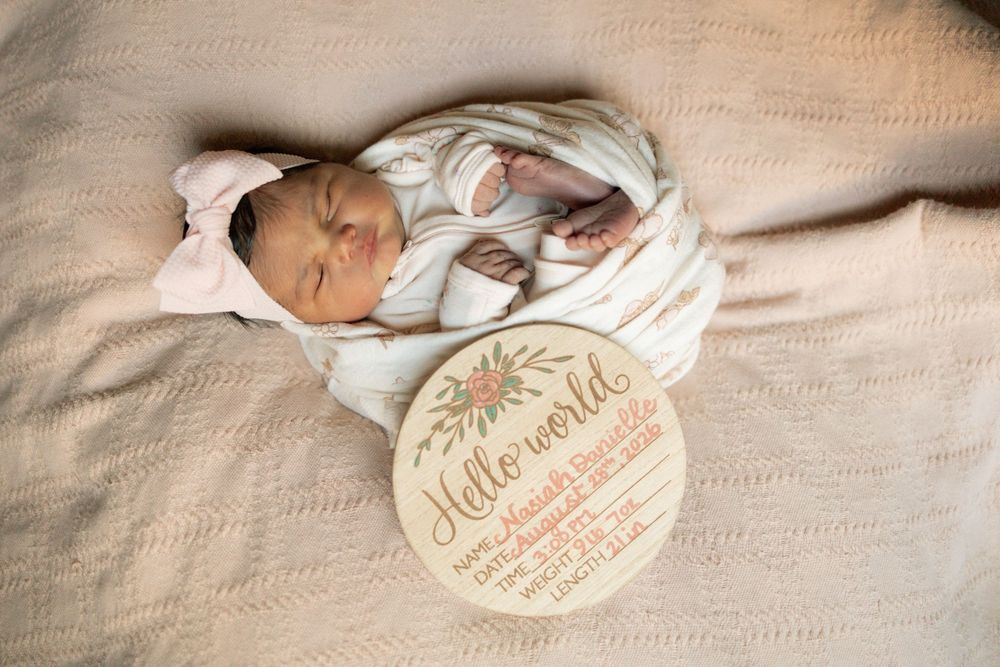 Newborn baby girl swaddled in white wrap laying beside a Hello World wooden birth stats announcement sign on beige blanket.