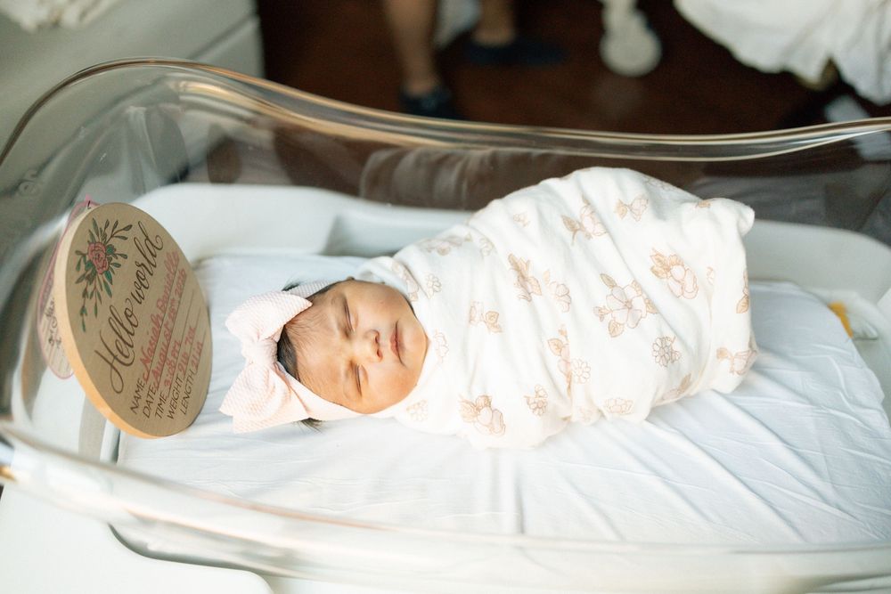 Newborn baby swaddled in floral wrap with pink hat sleeping peacefully in hospital bassinet.