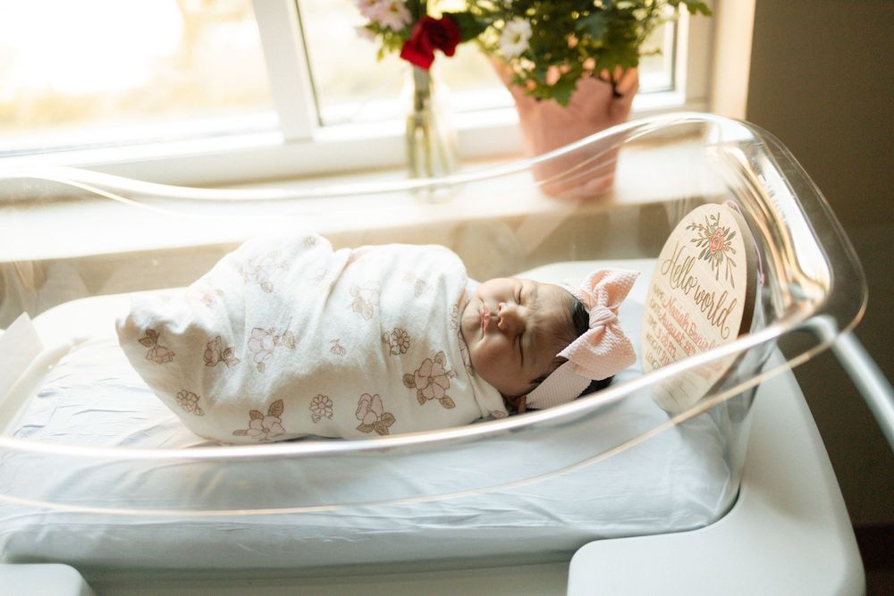 Newborn baby swaddled in white blanket sleeping peacefully in clear hospital bassinet near sunny window.