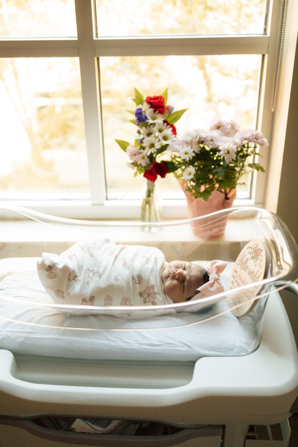 Newborn baby swaddled in hospital bassinet by sunny window with colorful flower bouquets.