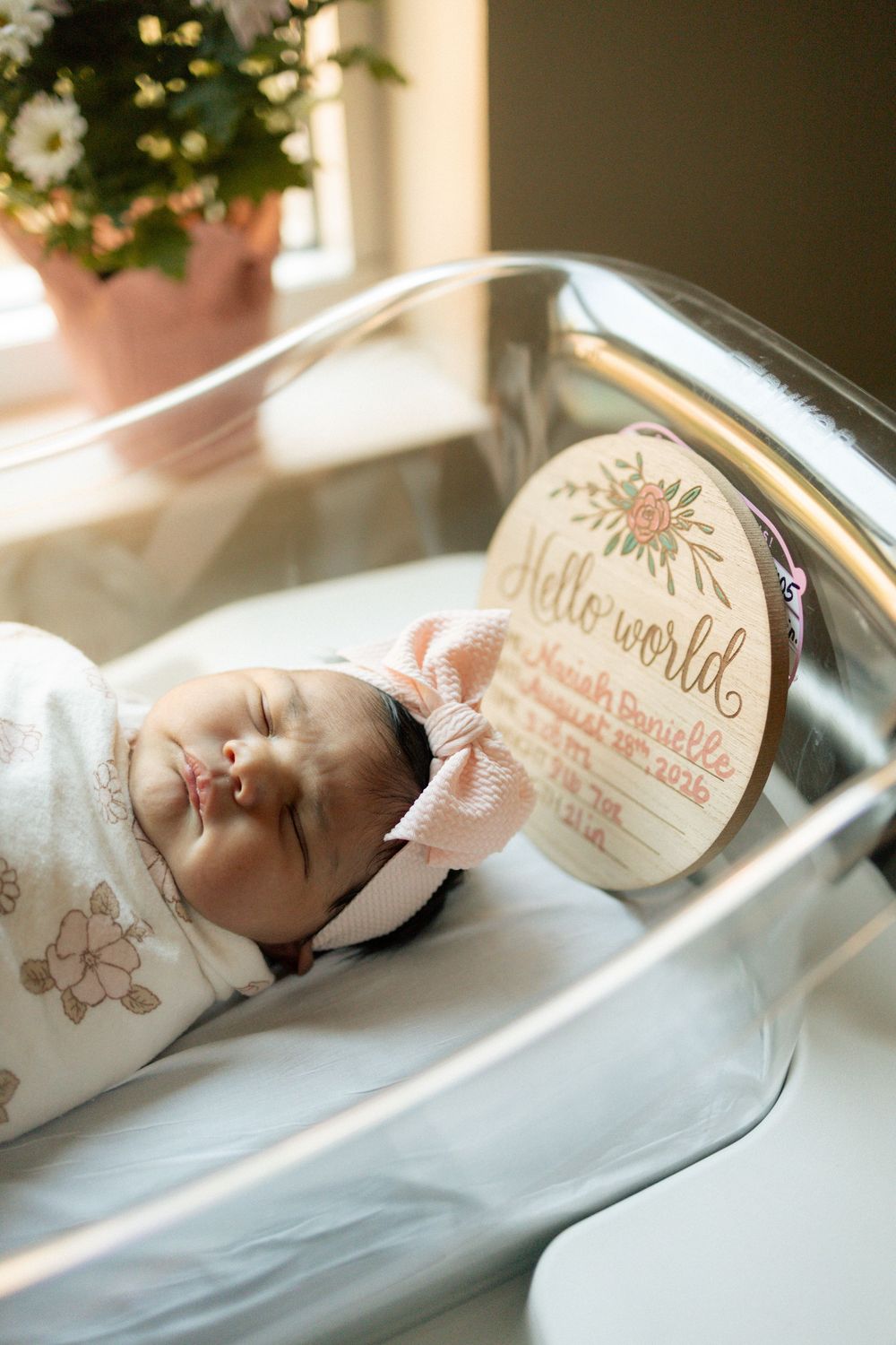 Newborn baby girl swaddled in floral wrap with pink bow headband, holding Hello World birth announcement sign in hospital bassinet.