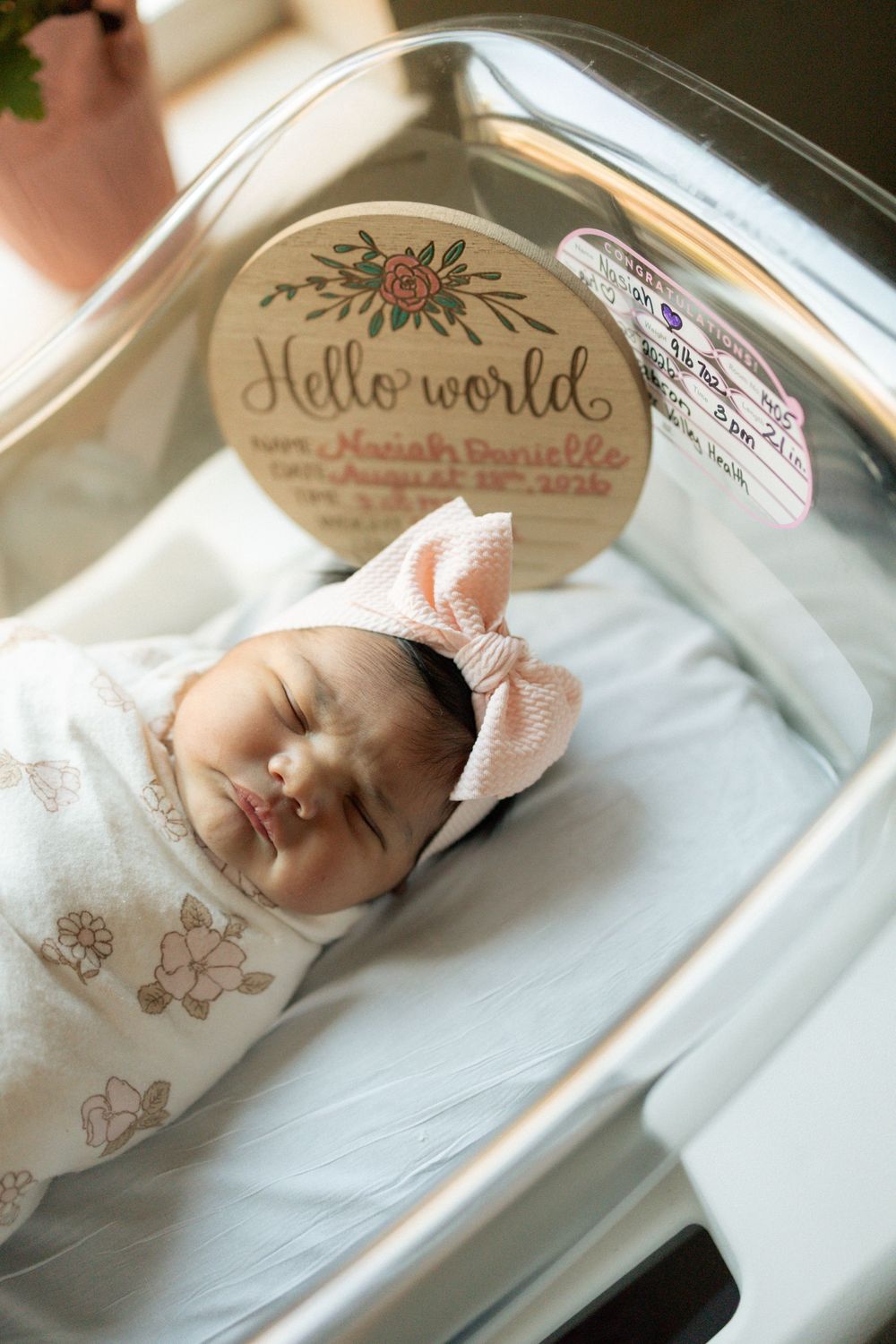 Newborn baby girl Naiah Danielle swaddled in floral wrap with pink bow headband in hospital bassinet with Hello World sign.