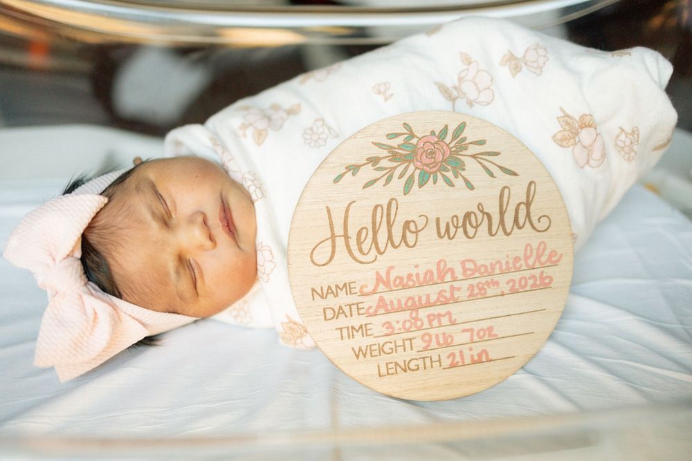 Newborn baby girl Mariah Danielle swaddled in floral wrap beside a wooden Hello World birth announcement sign.