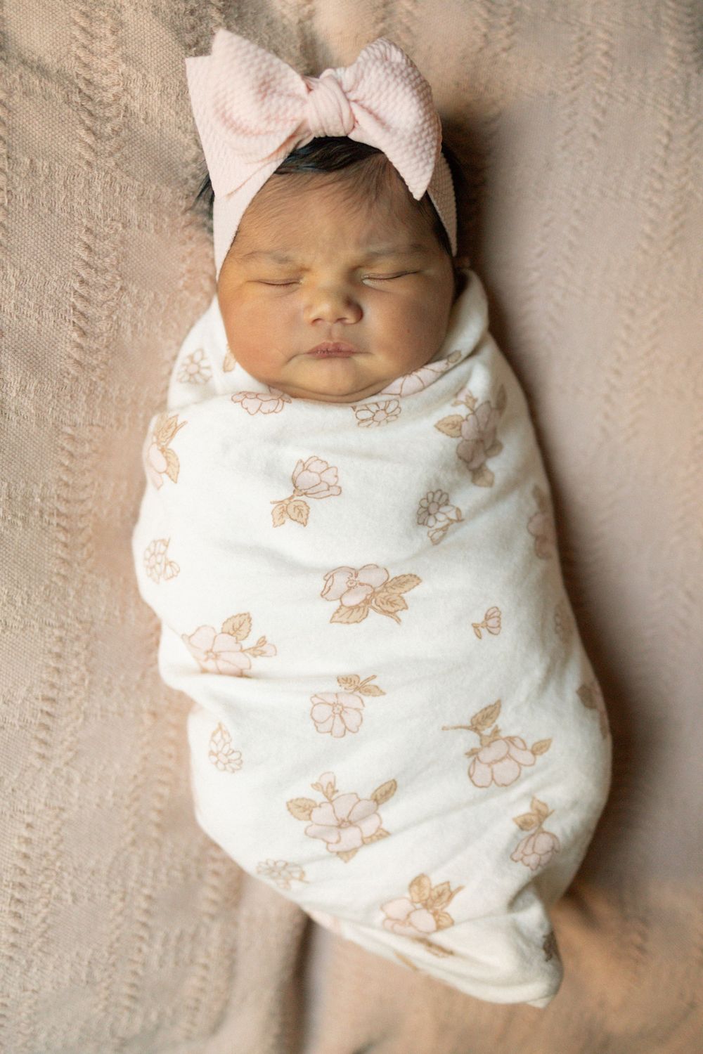 Newborn baby girl swaddled in white floral muslin wrap wearing pink bow headband, sleeping peacefully.