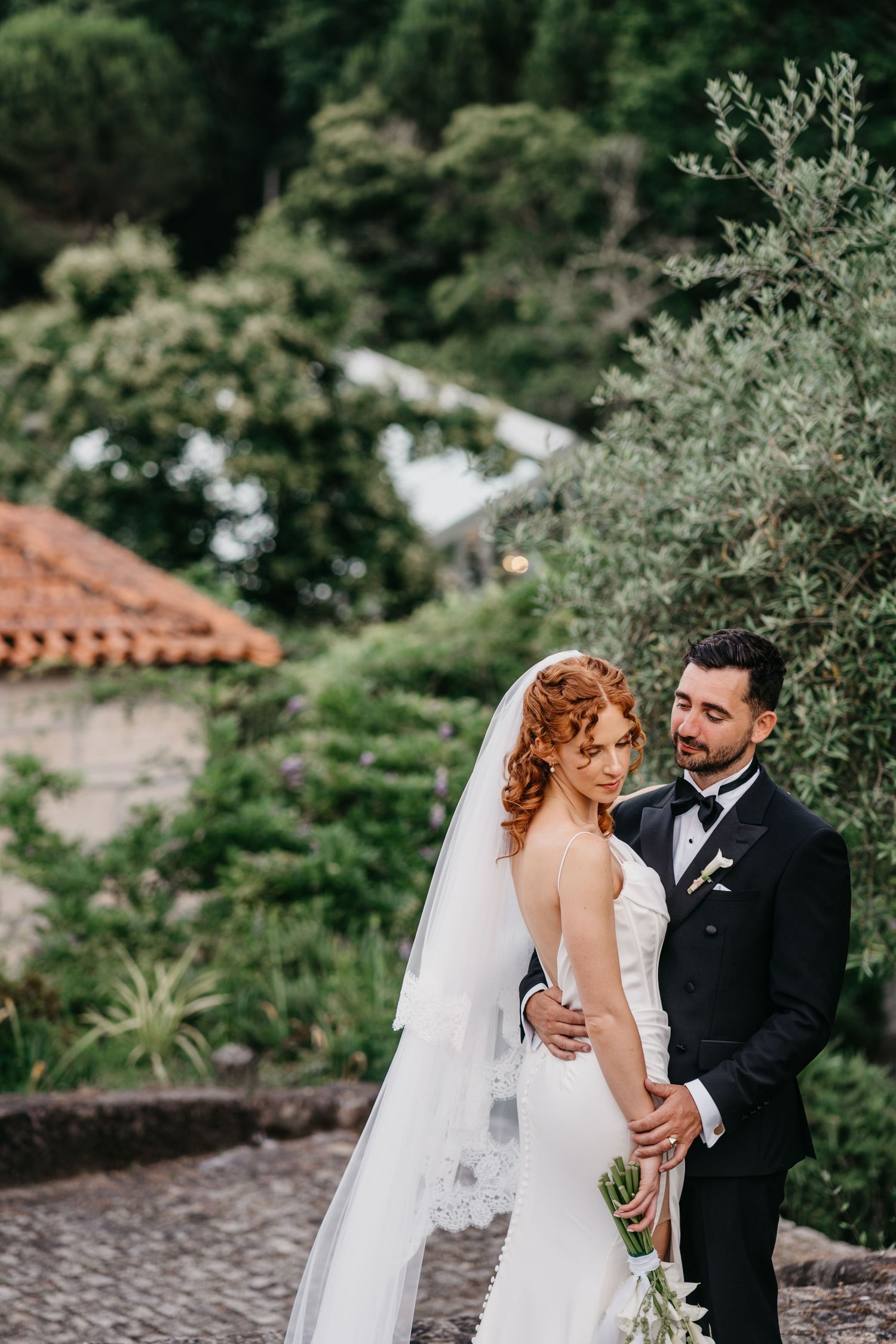 Bride in white gown and veil with groom in black tuxedo embracing outdoors amid lush greenery and terracotta rooftops.