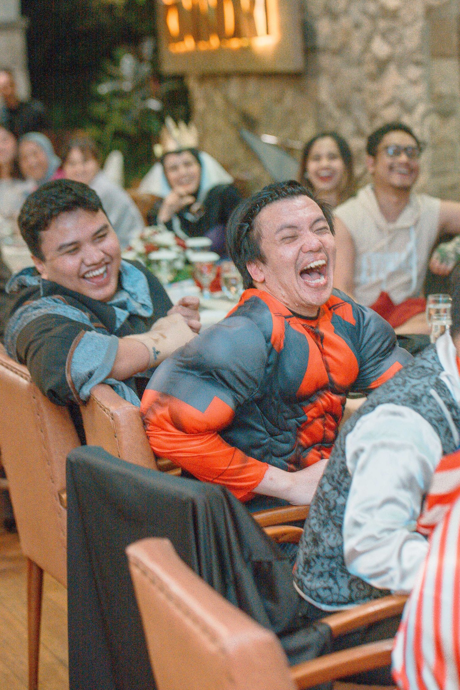 Two men in costumes laughing joyfully at a festive party event with guests seated at decorated tables.