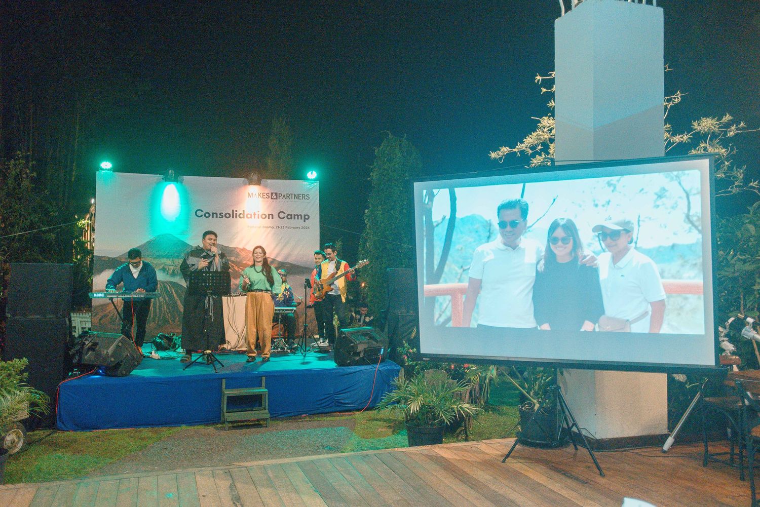 Outdoor evening concert with live band performing on stage next to a large projection screen at a Consultation Camp event.