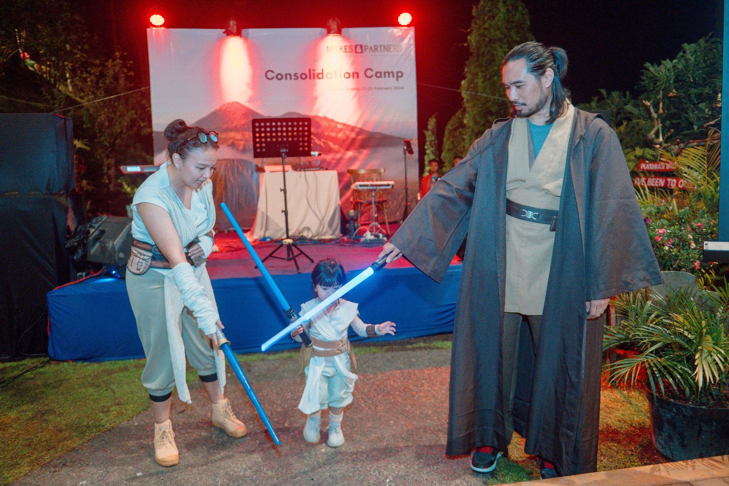 Two adults in Star Wars Jedi costumes with lightsabers play with a small child at an outdoor event.