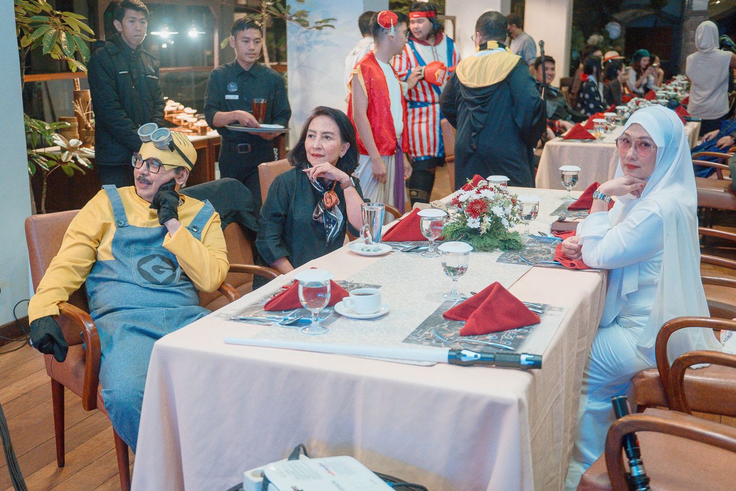 People in costumes at a festive dinner party, including a Minion outfit, clown, and blue character costume.