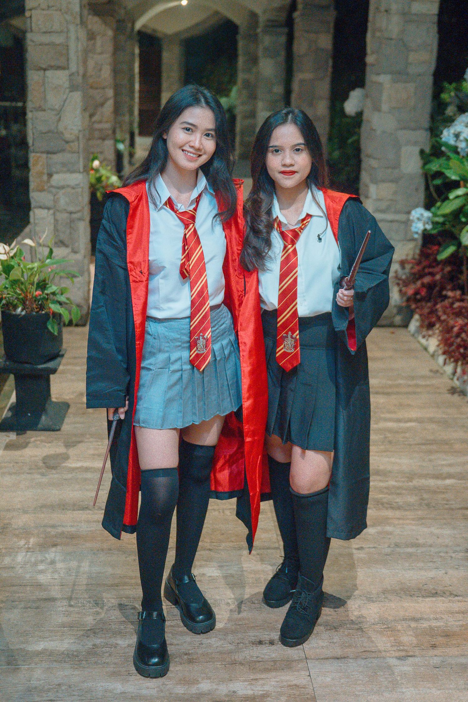 Two girls in Harry Potter Hogwarts uniforms with red ties, black robes, and wands pose together outdoors.