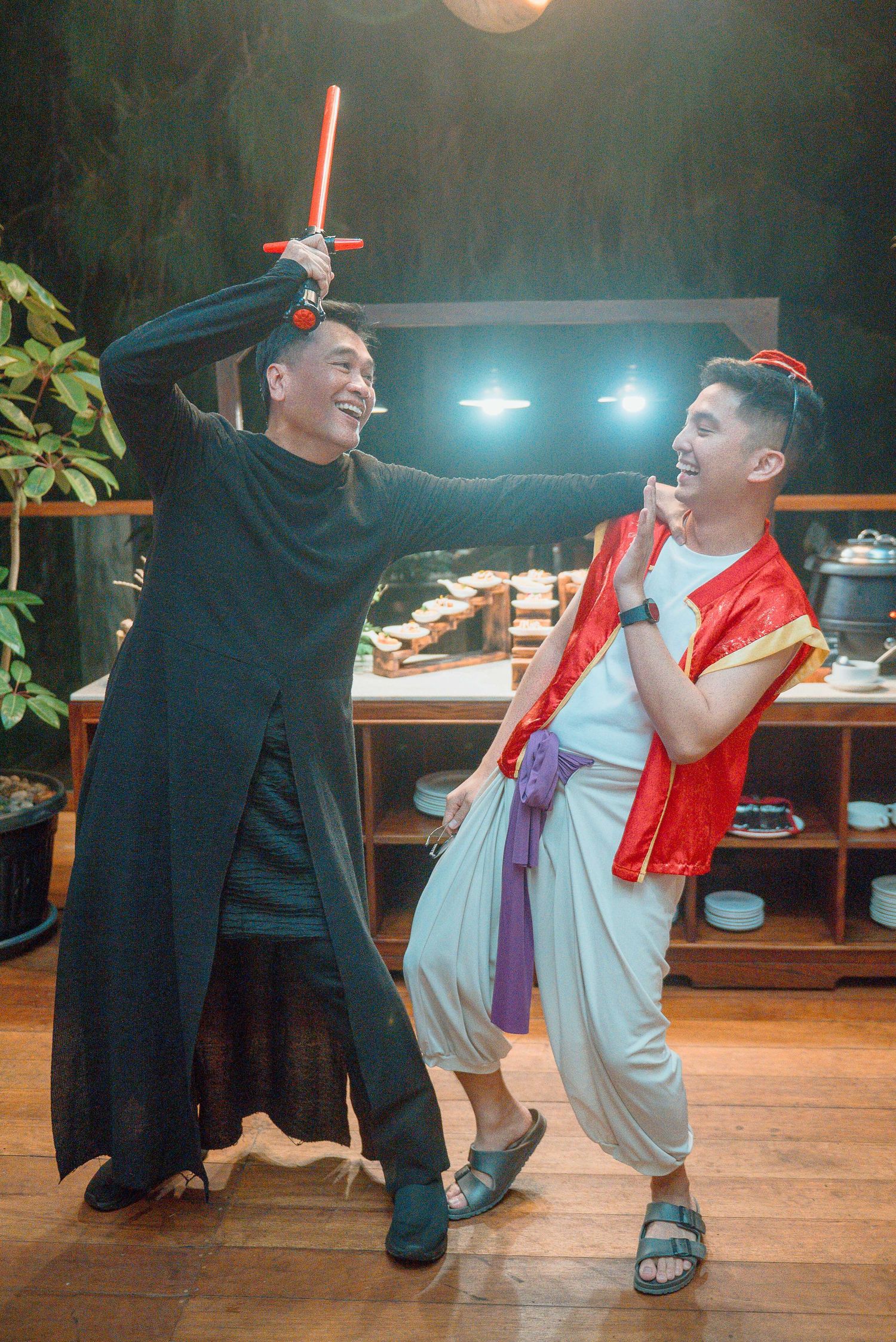 Two men in cosplay costumes pose playfully, one dressed as a Sith with a red lightsaber, the other as Aladdin.