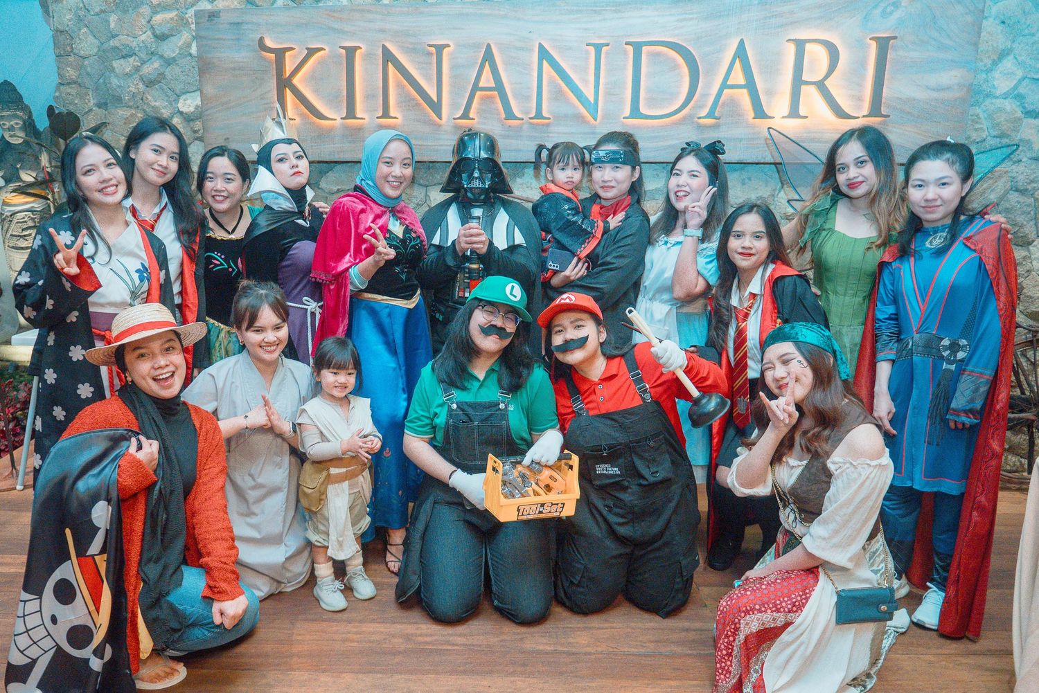 Group of people in colorful costumes posing in front of Kinandari sign, including Mario, Darth Vader, and fantasy characters.